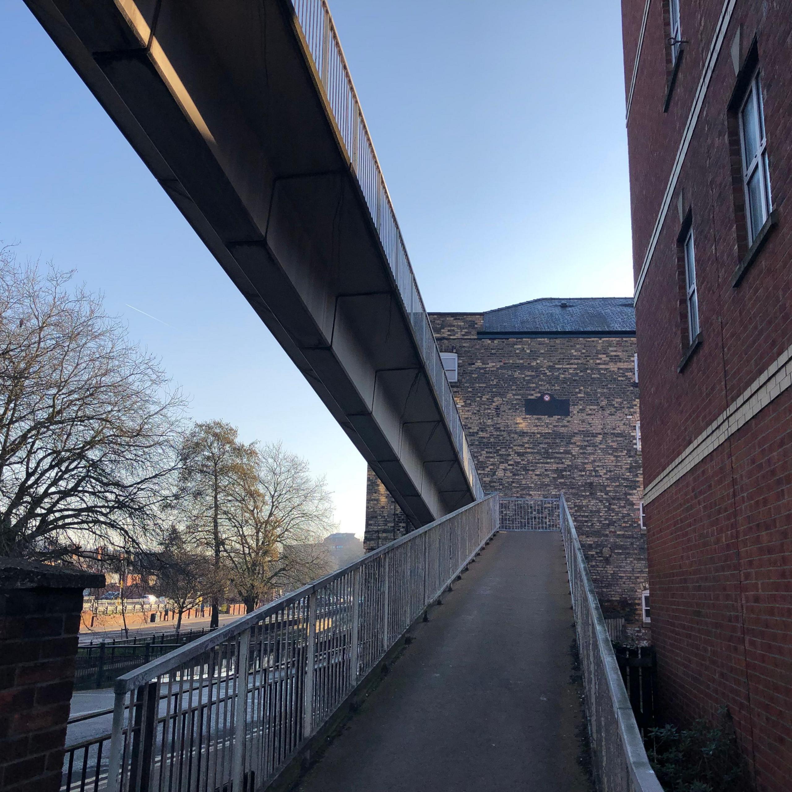 A view of a pedestrian bridge looking up a shallow slope from street level to a switch-back, with the main span crossing above. The bridge is concrete with metal railings.