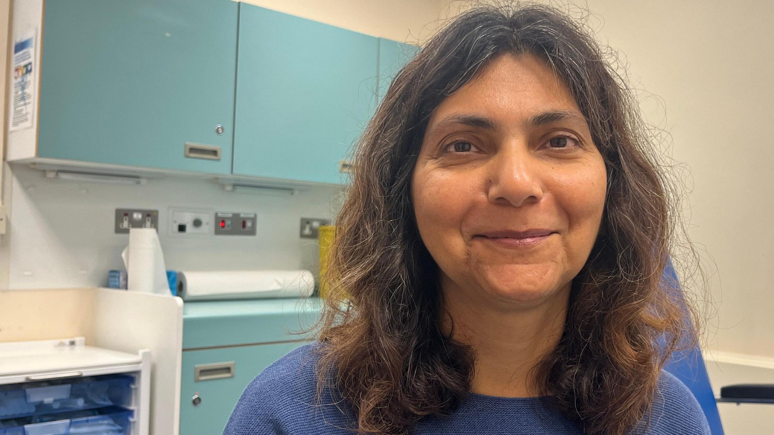 Pallavi Devulapalli, a woman who is sitting inside a doctors surgery consultation room. She has brunette shoulder length hair and is wearing a blue jumper. She is looking directly at the camera and smiling.