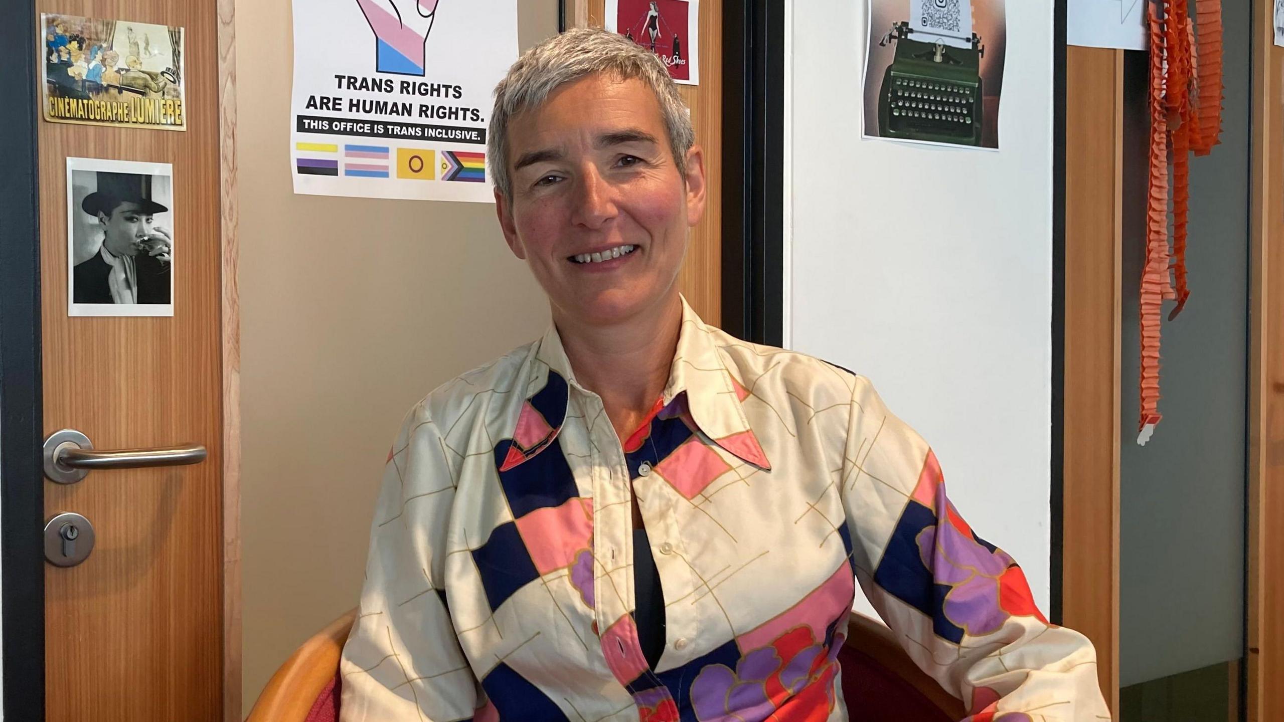 A woman with short grey hair and a cream-coloured blouse with some colourful patches sits in a chair, smiling at the camera. The wall behind her is decorated with pictures and posters, including one which reads: "TRANS RIGHTS ARE HUMAN RIGHTS".