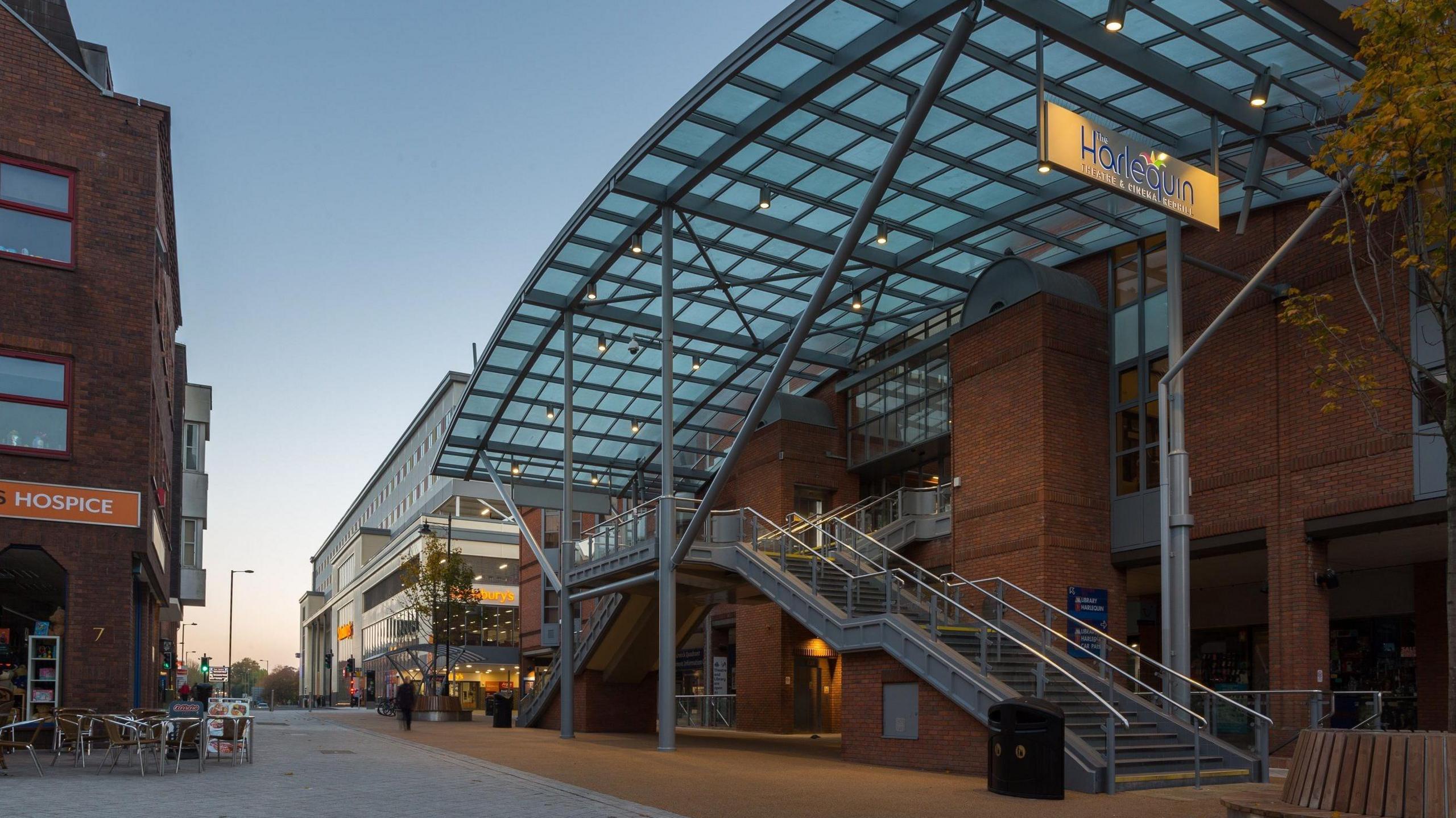 The outside of the Harlequin Theatre which is in a red brick building with a large glass canopy over the top and a sign hanging from it which reads Harlequin. In the background you can see a high street with a Sainsburys along the road