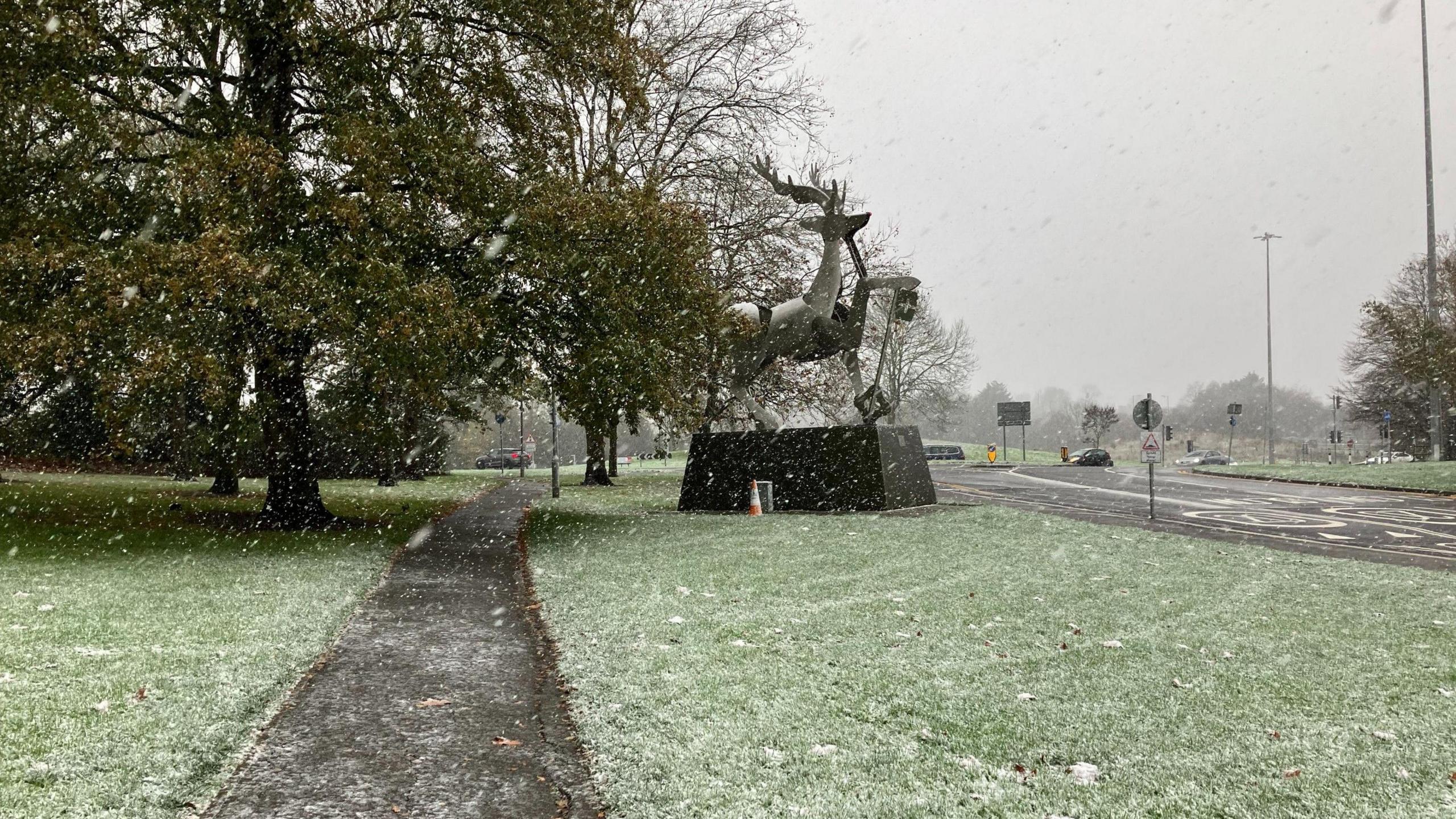 The stag statue on the University of Surrey campus, on a plinth in front of a tree, is seen through falling snow, with some snow beginning to lay on the grass around it.
