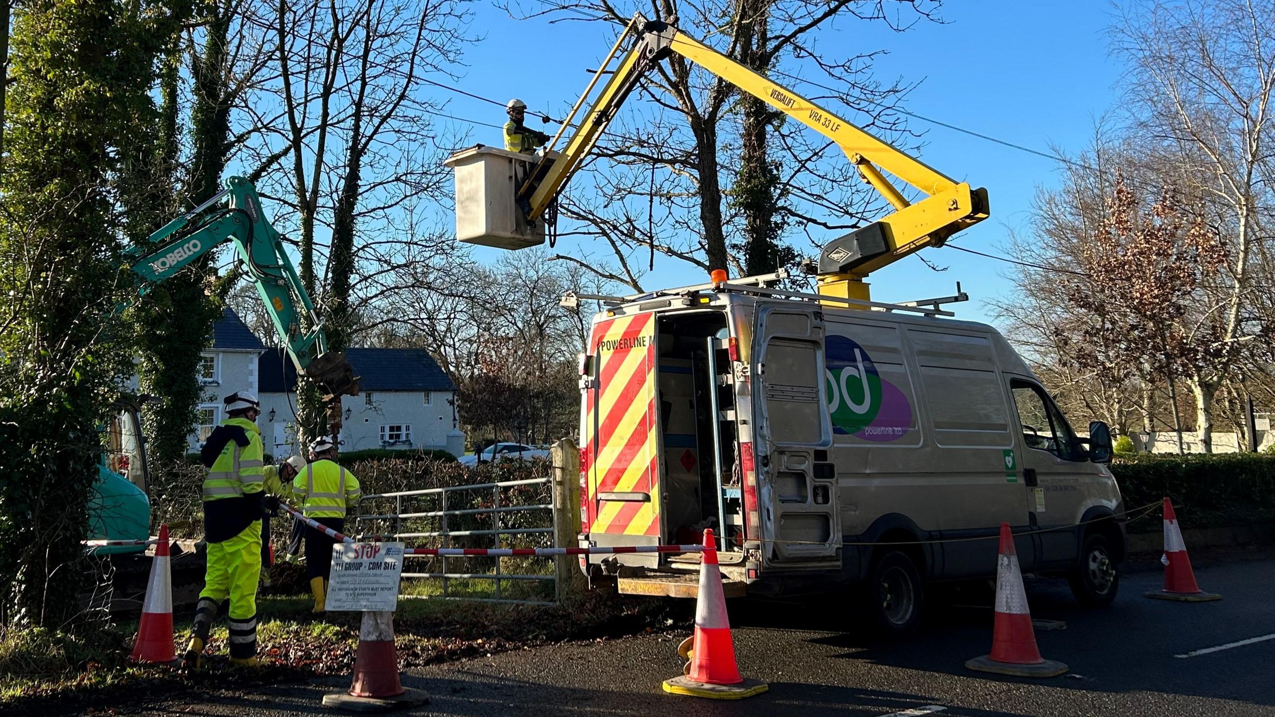 A man on a cherry-picker van near some power lines. The van is parked at the side of a road with some traffic cones around it. Tow other man in high-vis are standing off to the side. In the background some trees and a house.