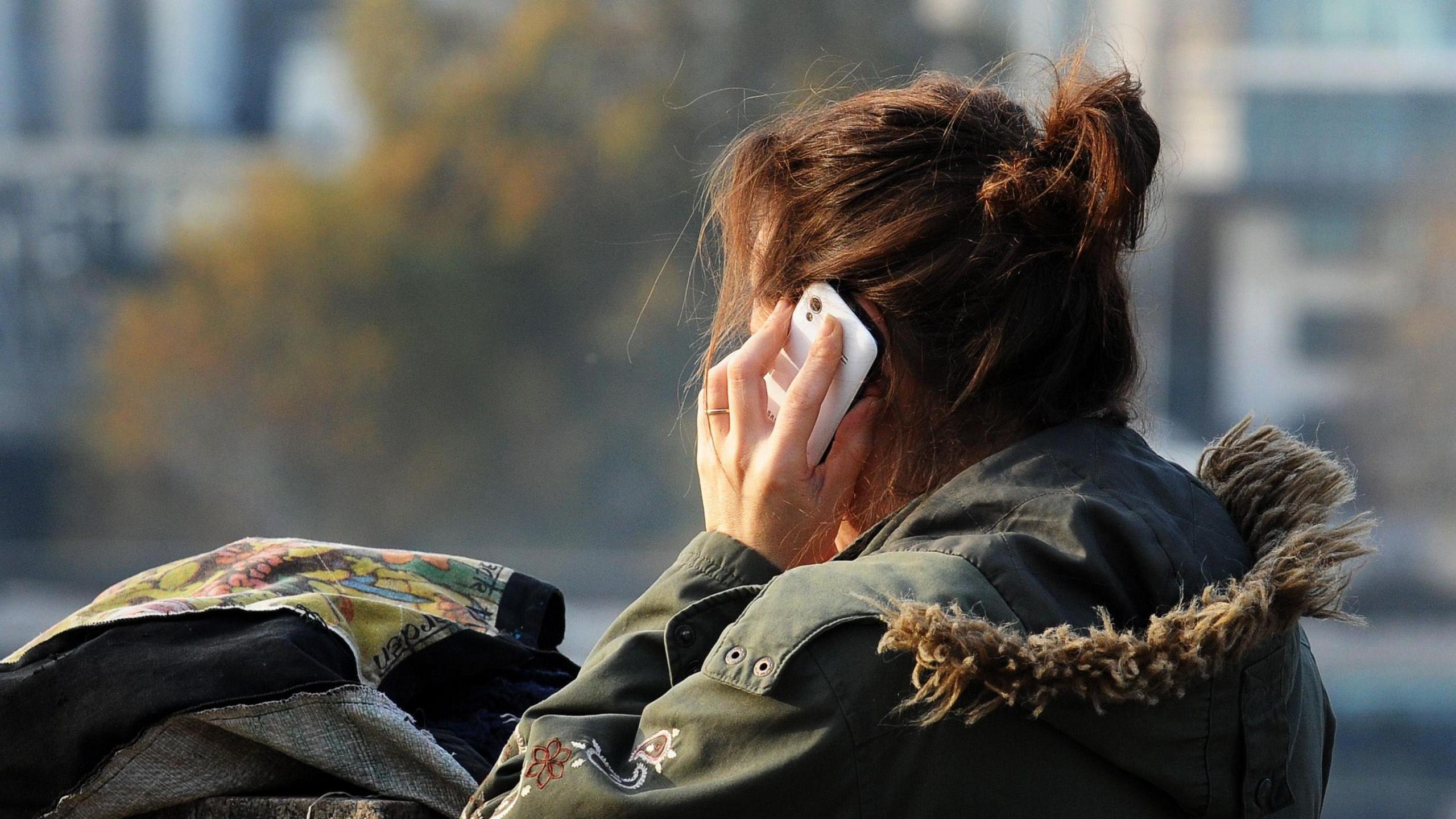 A woman is holding a mobile phone to her left ear. Her face is turned away from the camera. She is standing outside and has rested a bag on a wall in front of her. She has reddish brown hair in a loose bun and is wearing a coat with a hood that is trimmed with fur. She is wearing a ring on the ring finger of her left hand.