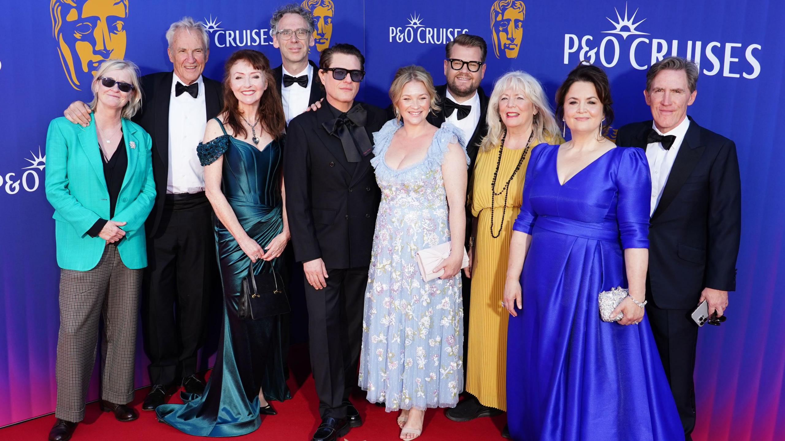 (Left to right) Christine Gernon, Larry Lamb, Melanie Walters, Robert Wilfort, Mathew Horne, Joanna Page, James Corden, Alison Steadman, Ruth Jones and Rob Brydon in evening dress as they attend the 2025 BAFTA Television Awards. They are pictured on a red carpet with a P&O cruises blue and purple advertising hoarding behind them.