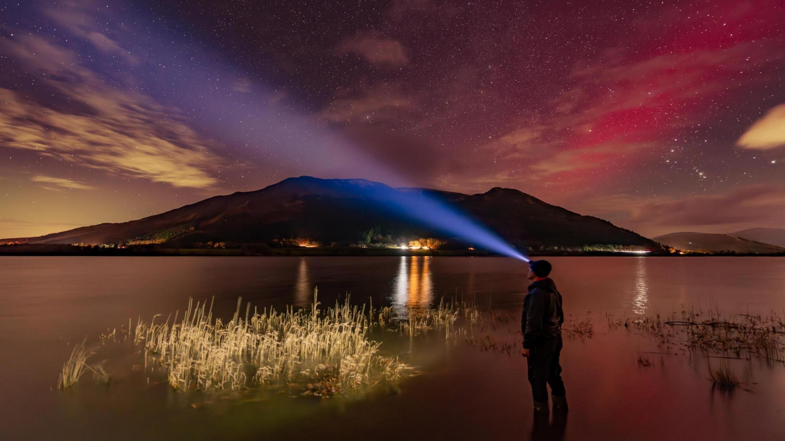 The Northern Lights are seen above a mountain. In the foreground, the man is illuminated by the light of his head torch.