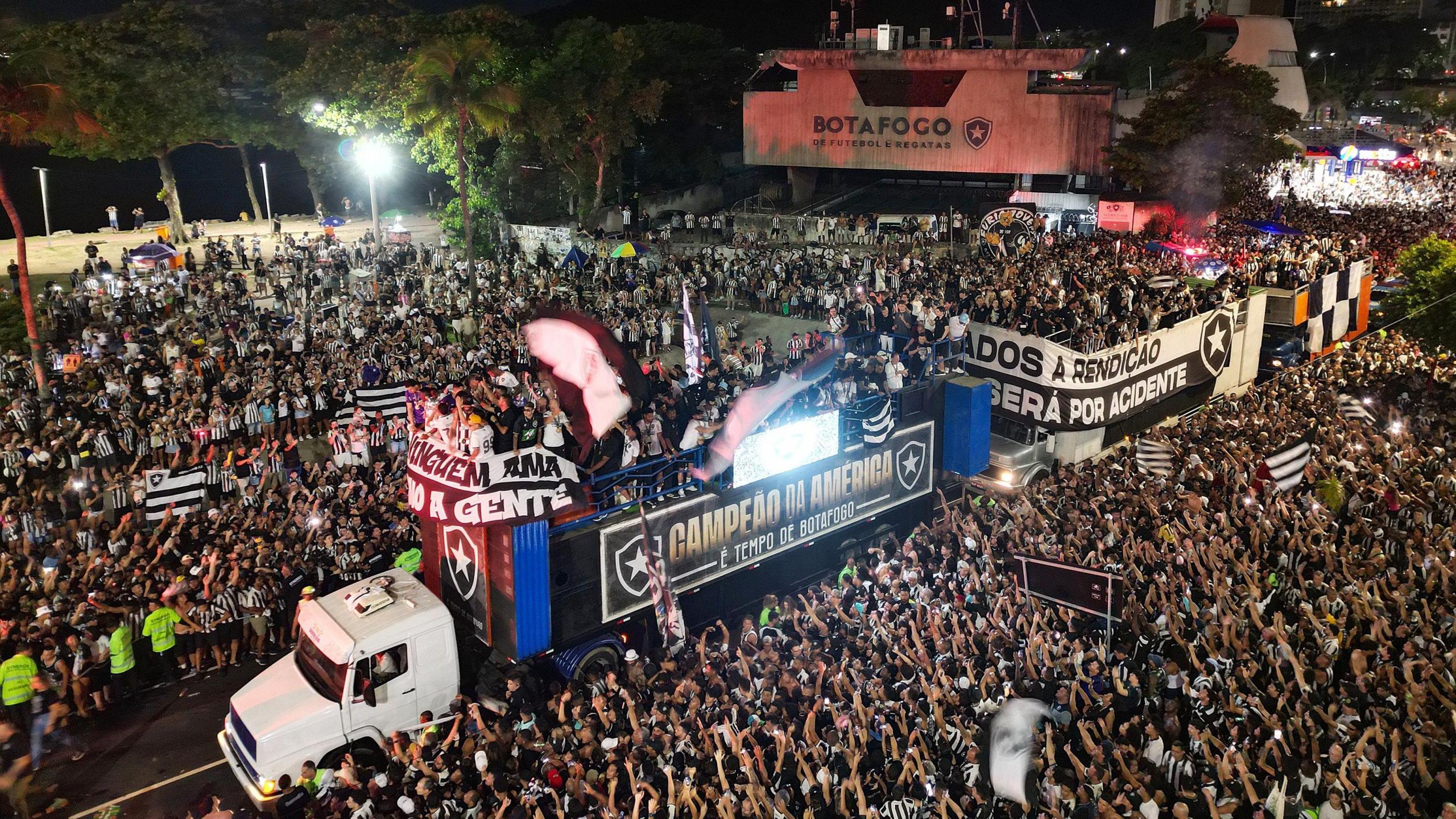 An articulated lorry, followed by a bus bearing Botafogo players, weaves its way through assembled fans gathered at a beach for a trophy celebration