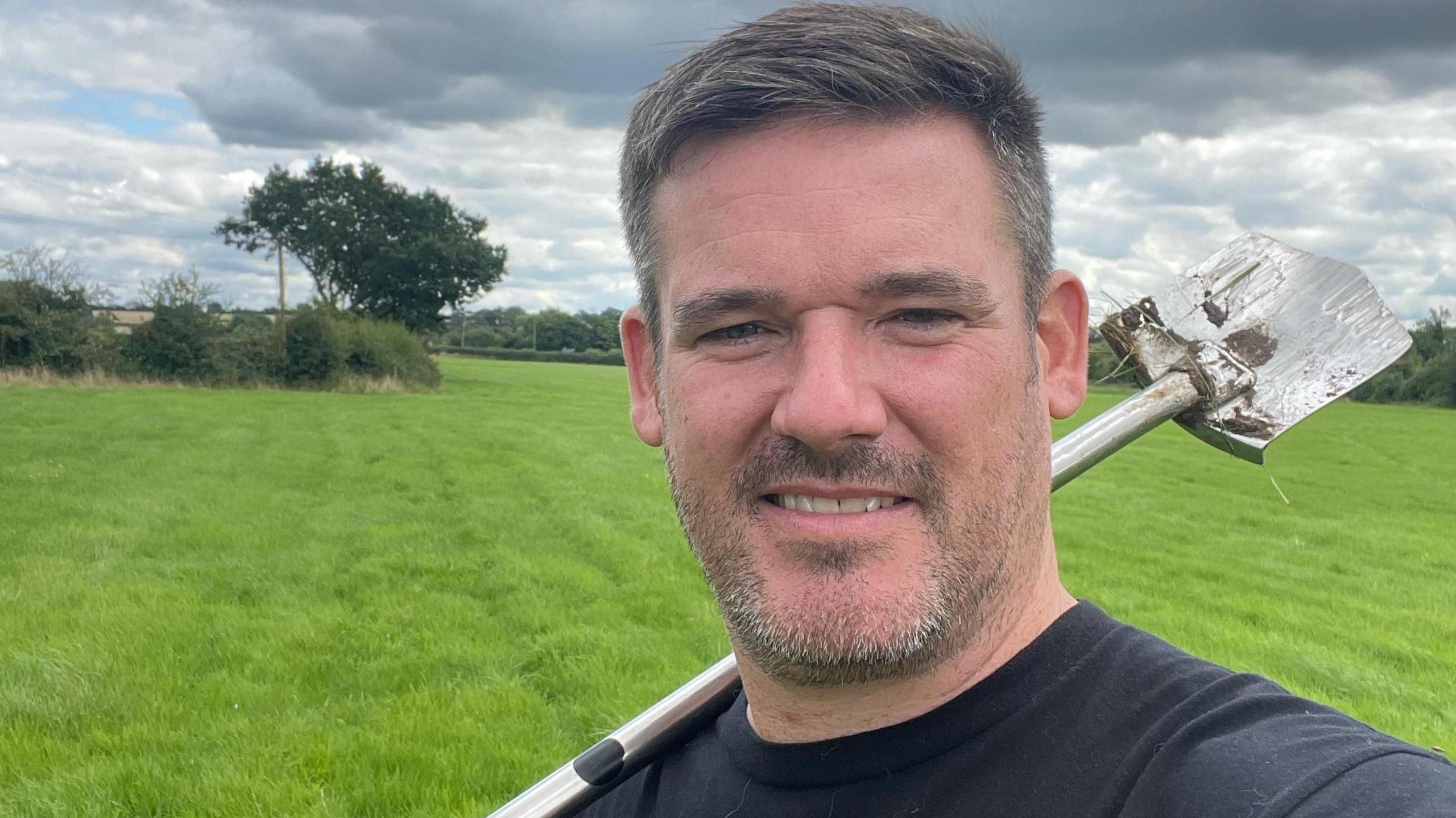 David Dunn with a shovel over his shoulder in front of a grassy field