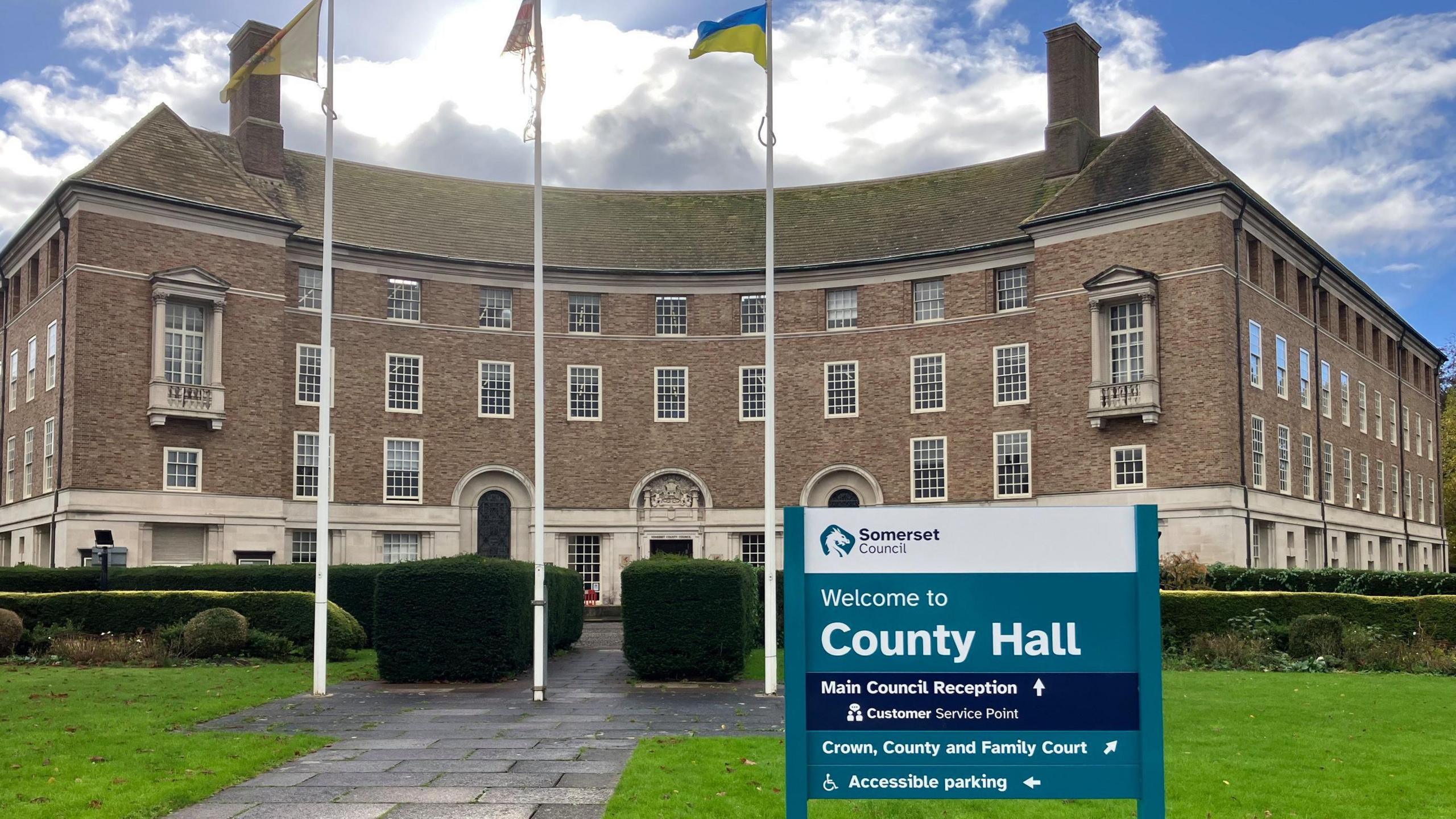 A sign reading 'welcome to county hall' outside a municipal building with flags flying