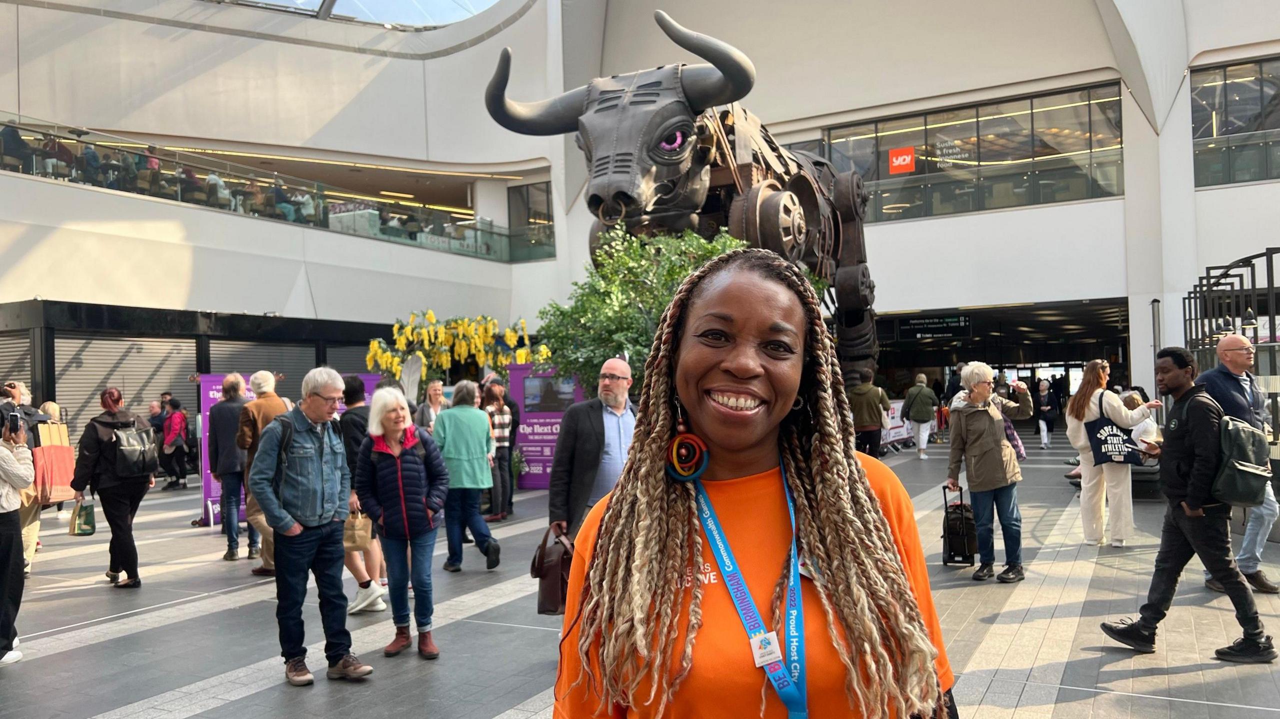 Maureen Levermore in a orange t-shirt. She is smiling and has blonde braids with black roots. She is standing inside Birmingham New street station.