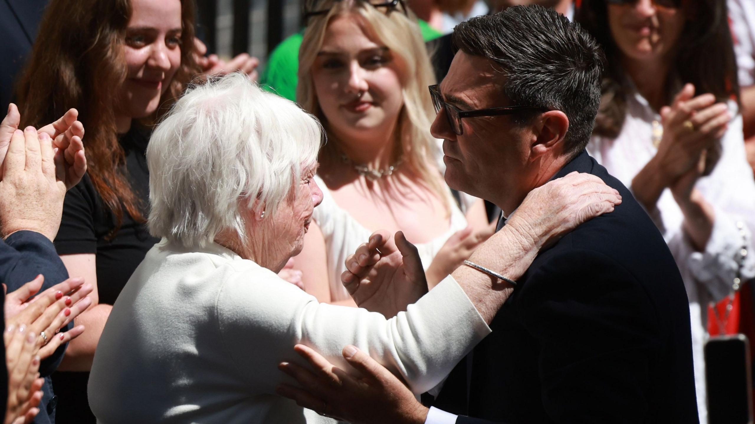 Andy Burham, side-on, is holding the elbow of his mother Eileen, who has grey hair and is wearing a white jumper. They are about to embrace.