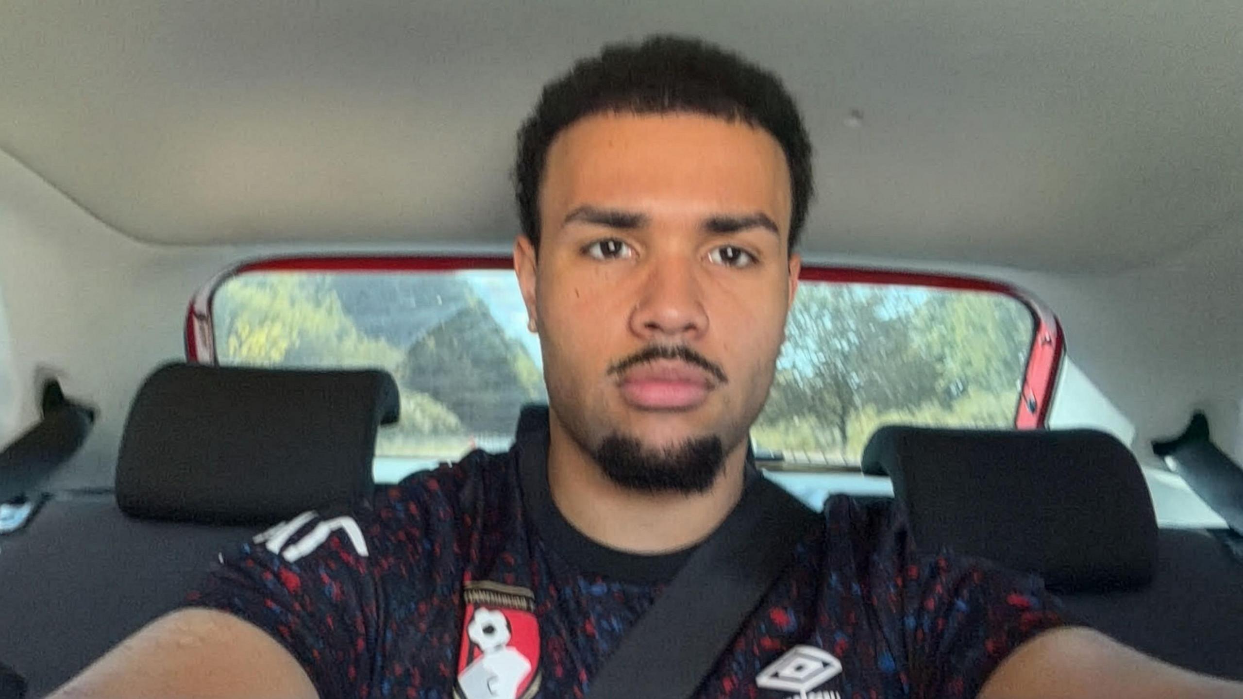 A man with short black hair is wearing a football shirt and sitting in the back of a car. 