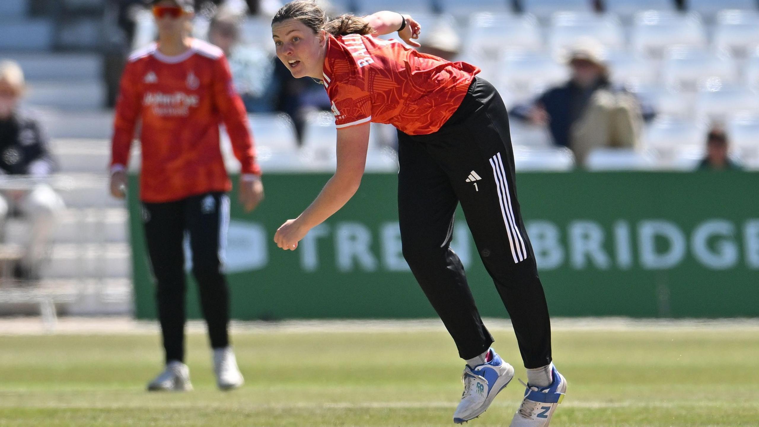 Grace Ballinger in bowling follow-through at Trent Bridge, wearing 2026 Blaze Metro Bank One-Day Cup kit (orange with fine detailing of the old and new pavillion.)
