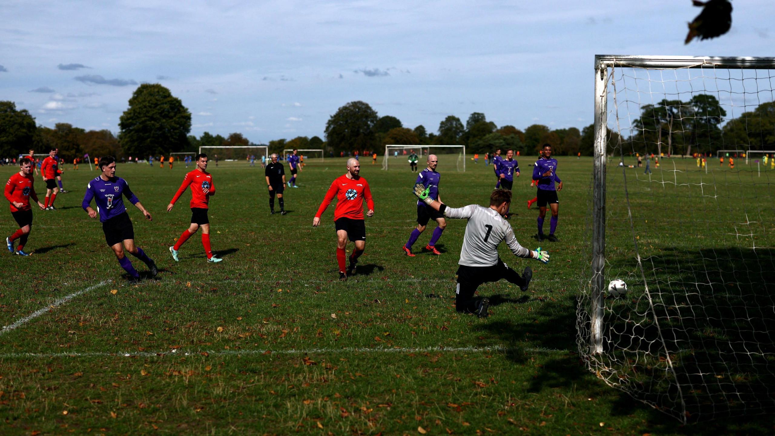 A player in a dark purple top and black shorts shoots a football across a goalkeeper in a white top with black shorts and towards a goal with a net. In the shot are other players from the shooter's team and their opponents, who are in orange tops and black shorts. The game is played as part of the Downs League in Bristol and several other matches can be seen going on in the background, along with a line of trees.