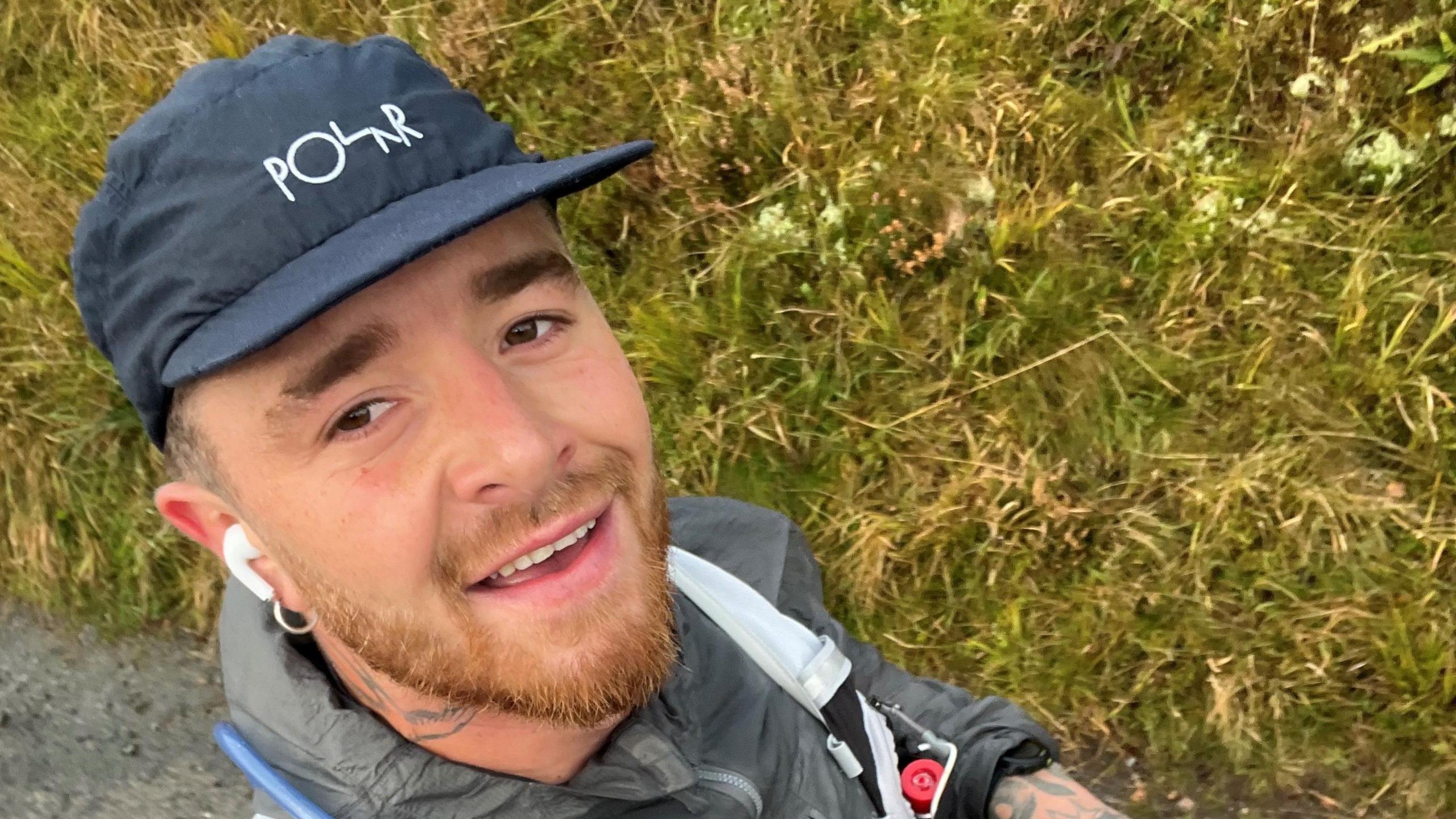 Jonah Evans, wearing a grey cap and in-ear headphones walking besides some grass