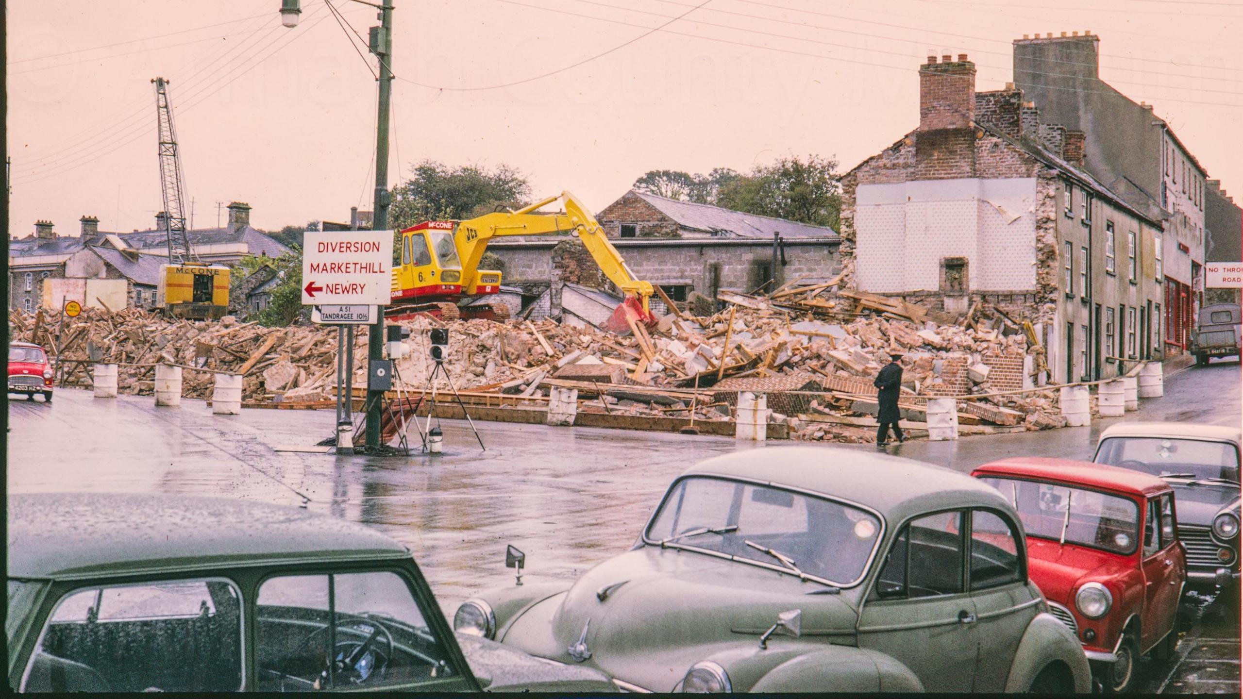 A digitalised photo from a film of wreckage from a demolition. Old cars are parked along the street. 
Looking south at the demolition of the buildings on the corner of Newry Road and Barrack Street. The jail can be seen on the left and Palace Row on the right, a crane and JCB digger with the name McCone sit on top of the demolished rubble.