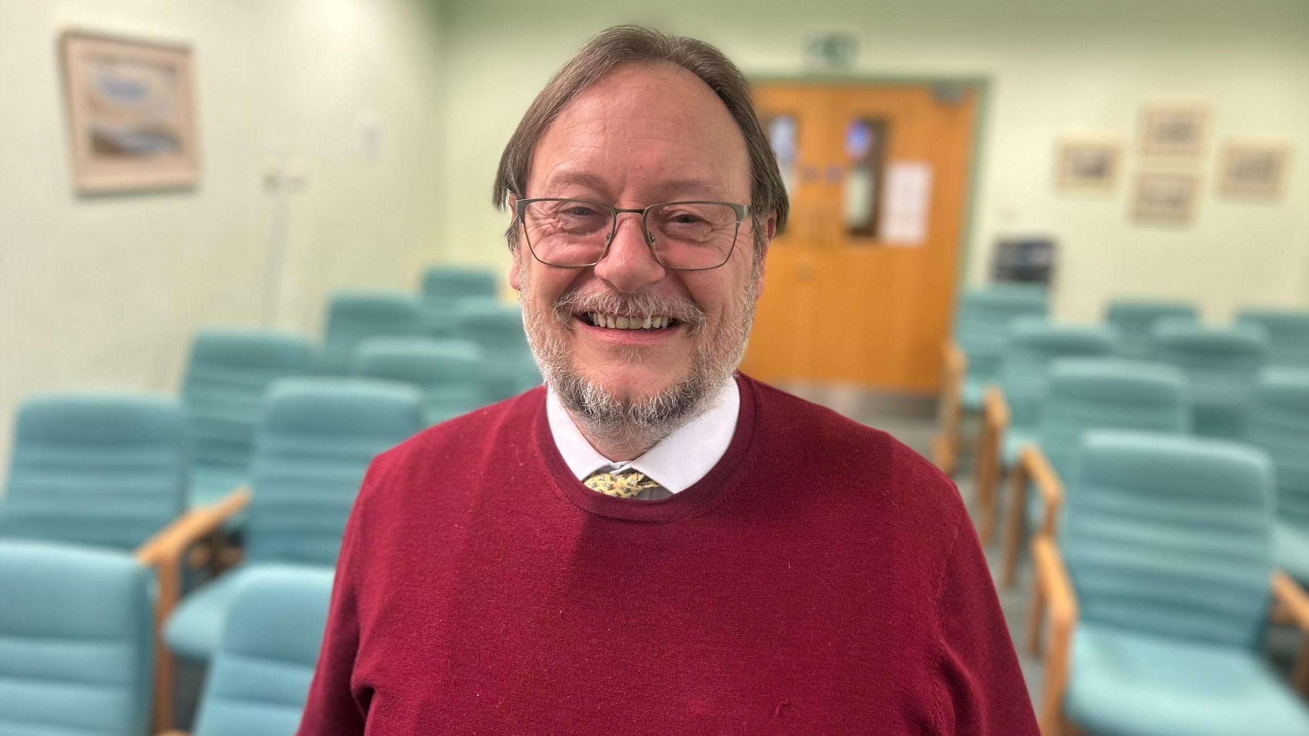 Phil Banfield smiling. He is wearing a red jumper and glasses and has short brown hair and grey beard. there are teal green chairs in the background.