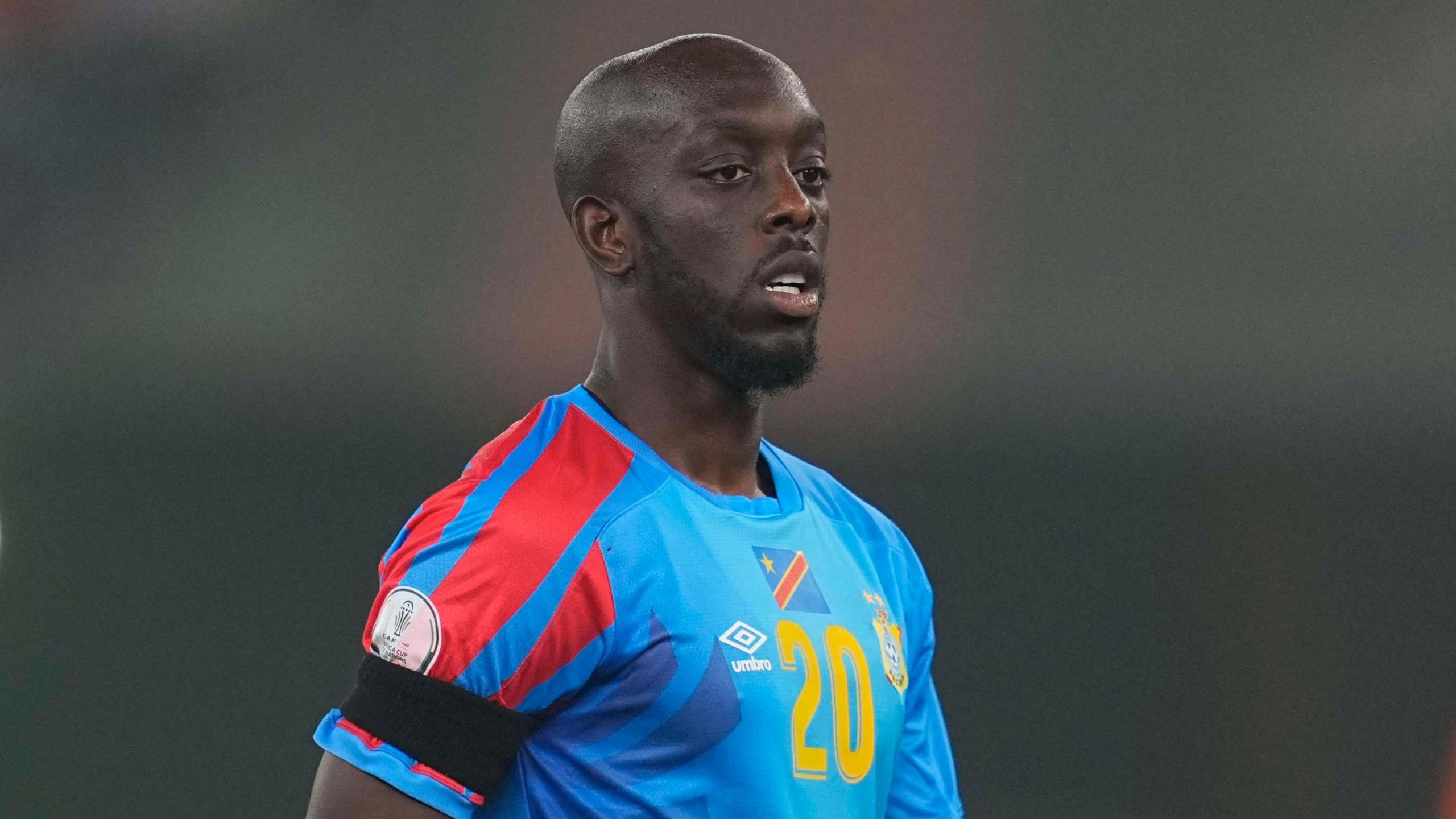 Yoane Wissa is seen from waist up wearing a blue DR Congo shirt with a national flag above the number 20 on the chest and red detail on the right shoulder. He has a black armband on his left upper arm, while he also sports a black beard and shaved head