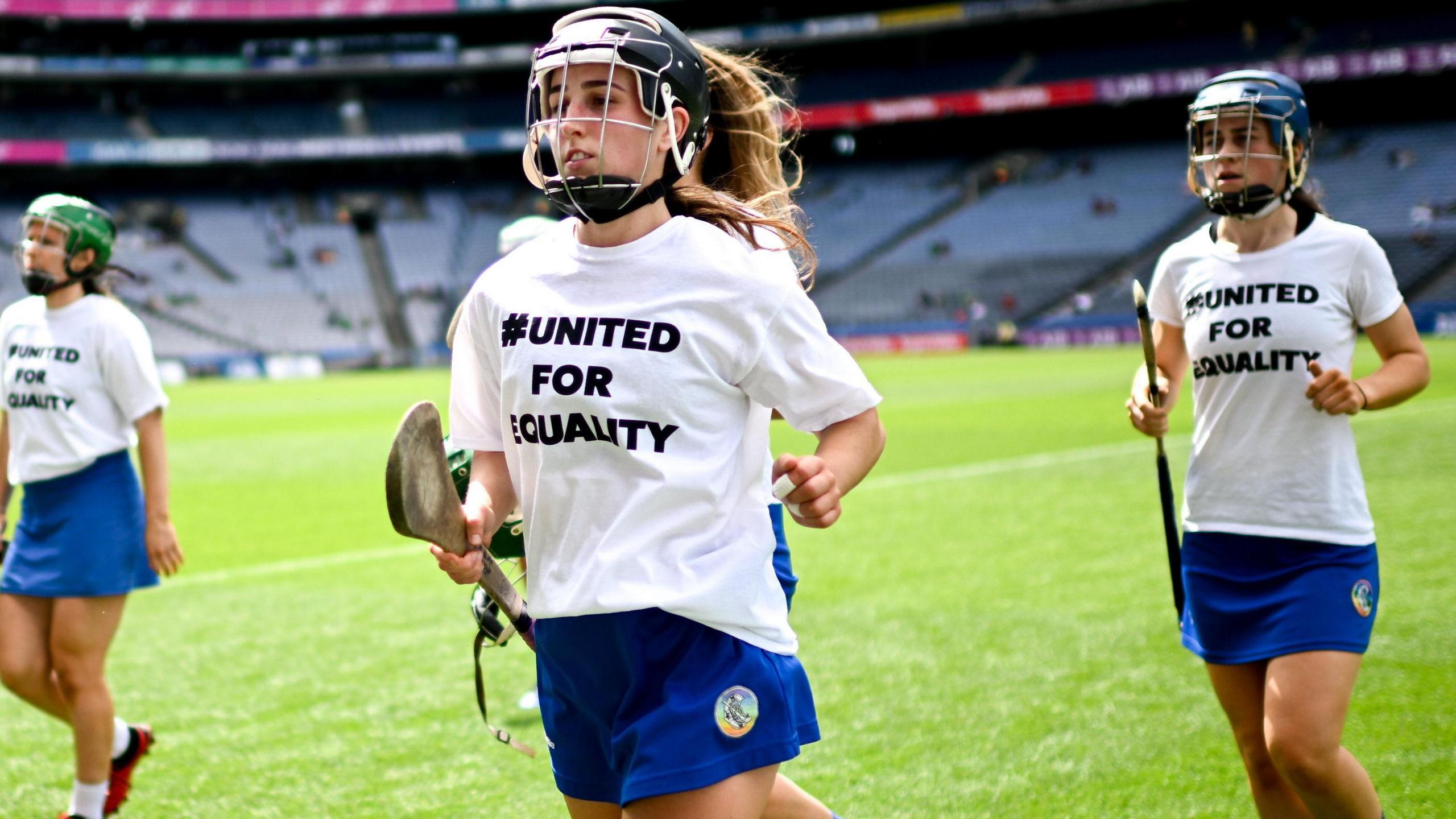 Three women jog off a pitch in a stadium. They are each wearing a helmet and hold a camogie hurley. They are wearing matching white t shirts that say '#united for equality'.