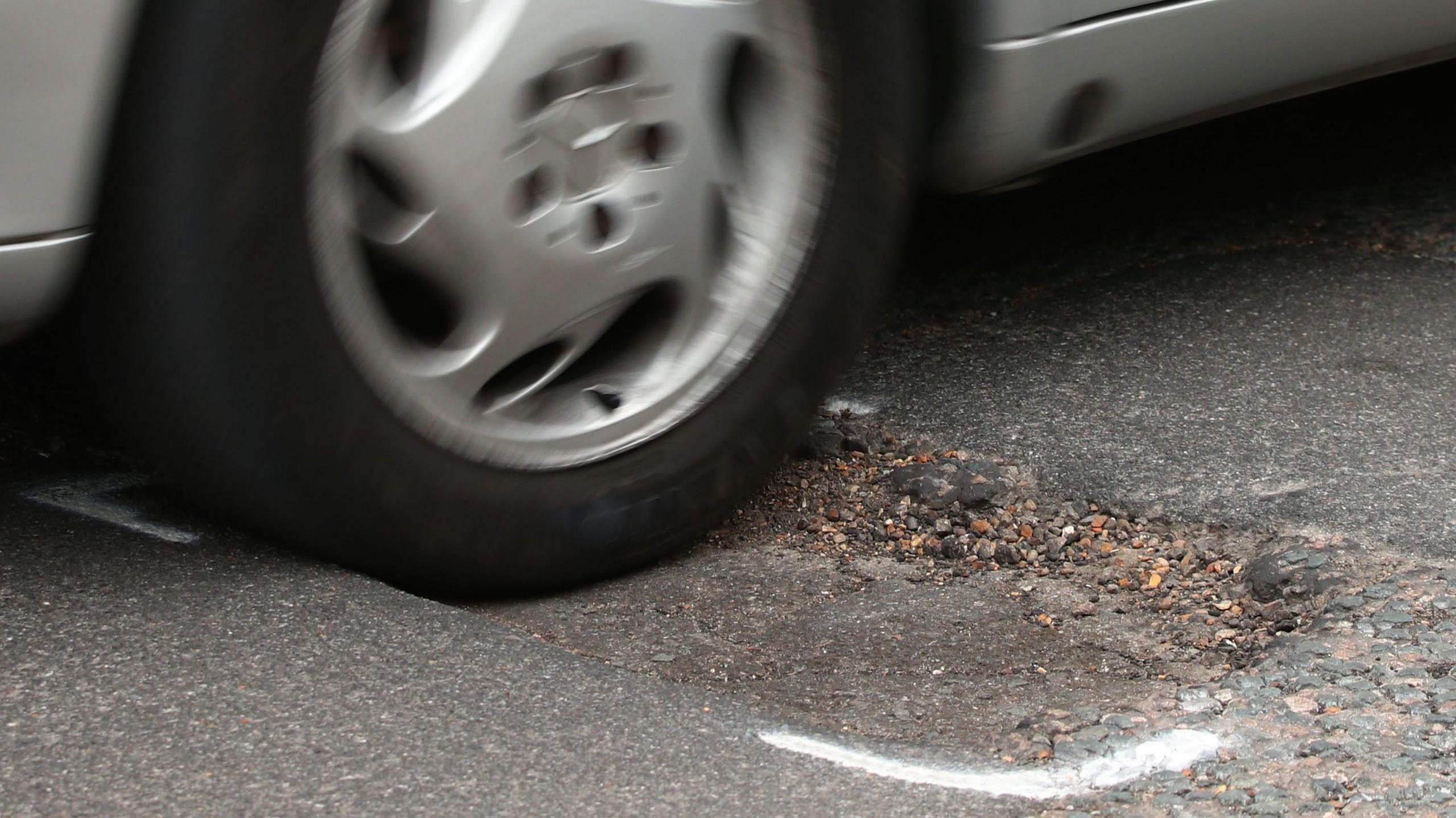 A silver car driving over a pothole on the road that has white spray paint markings around it.