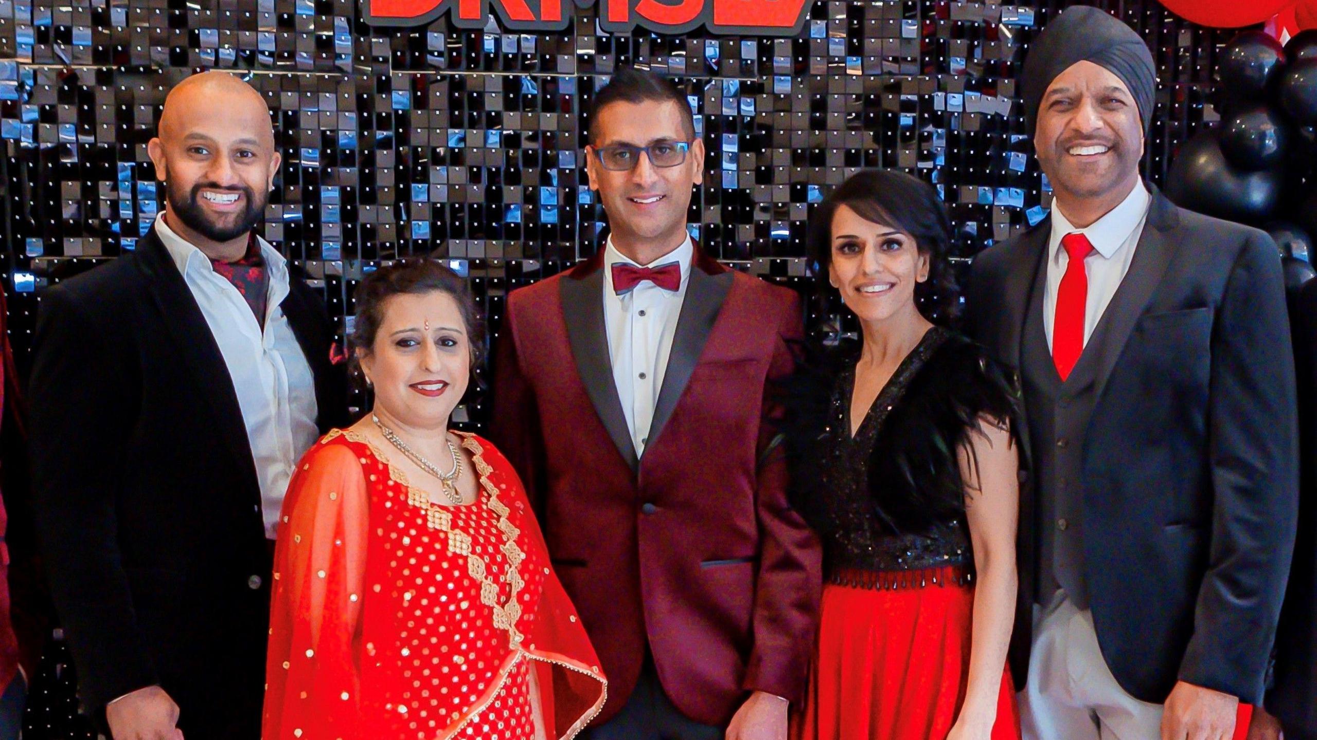 Five people standing in front of a large black shiny backdrop. There are three men and two women. 