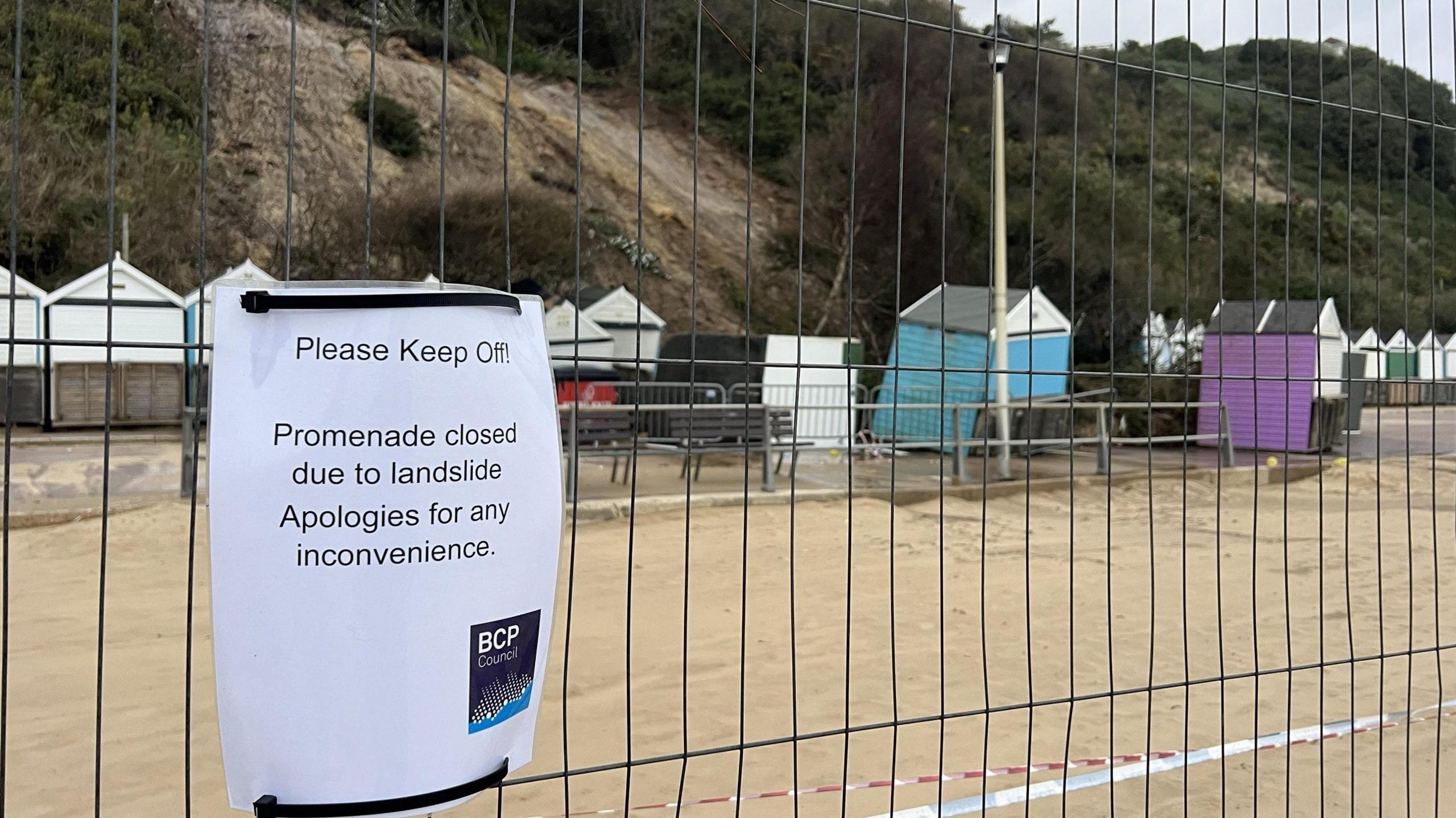 A sign on a fence with broken beach huts behind and a landslip behind