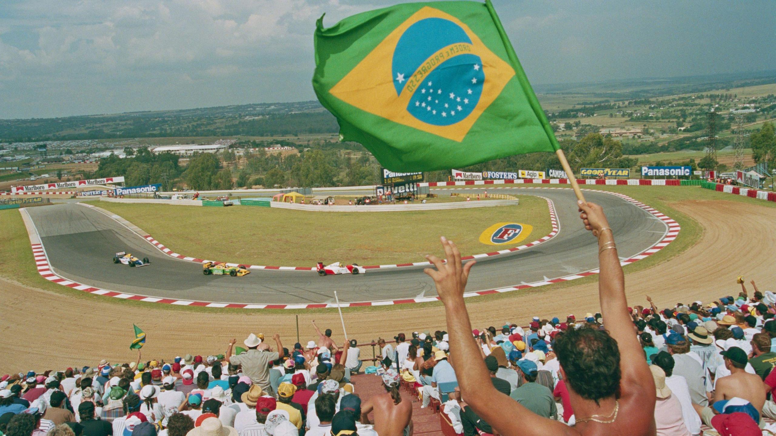 A topless man in a packed grandstand faces away from camera and waves a Brazilian flag as, in the background, three Formula one cars round a bend at the Kyalami track in South Africa