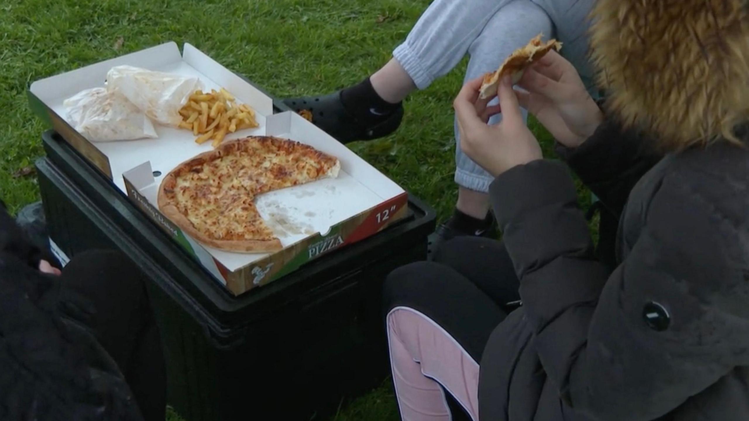 Young people eating pizza and chips at a youth session under a gazebo in a park. The food, in an open pizza box, rests on a box on wet grass. The young people wear coats and their faces are not shown.