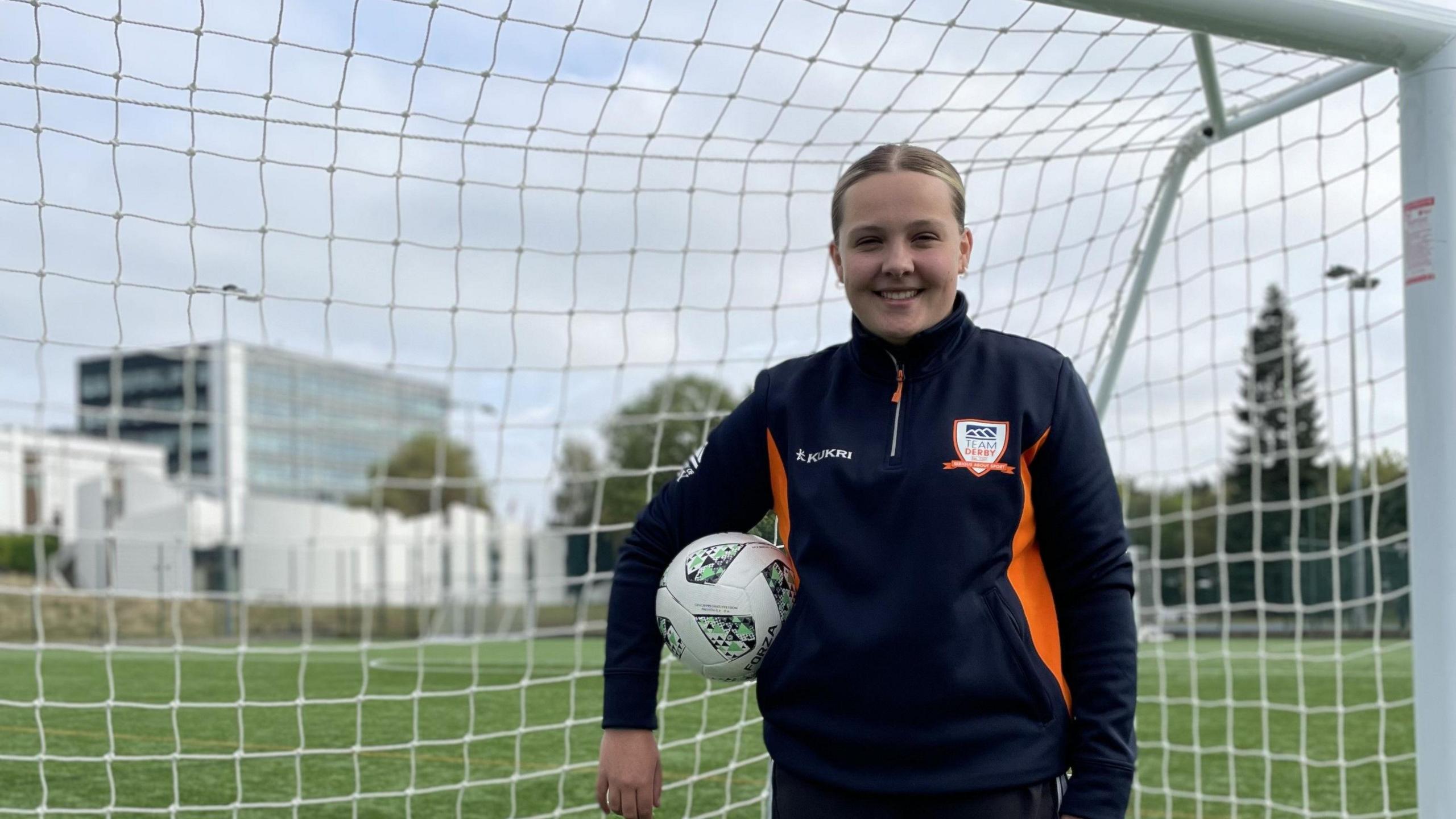 Ella Brennan standing with a ball under her arm, in front of a goal at the University of Derby's sports pitches. She is wearing a blue University of Derby jumper and has her hair tied back.