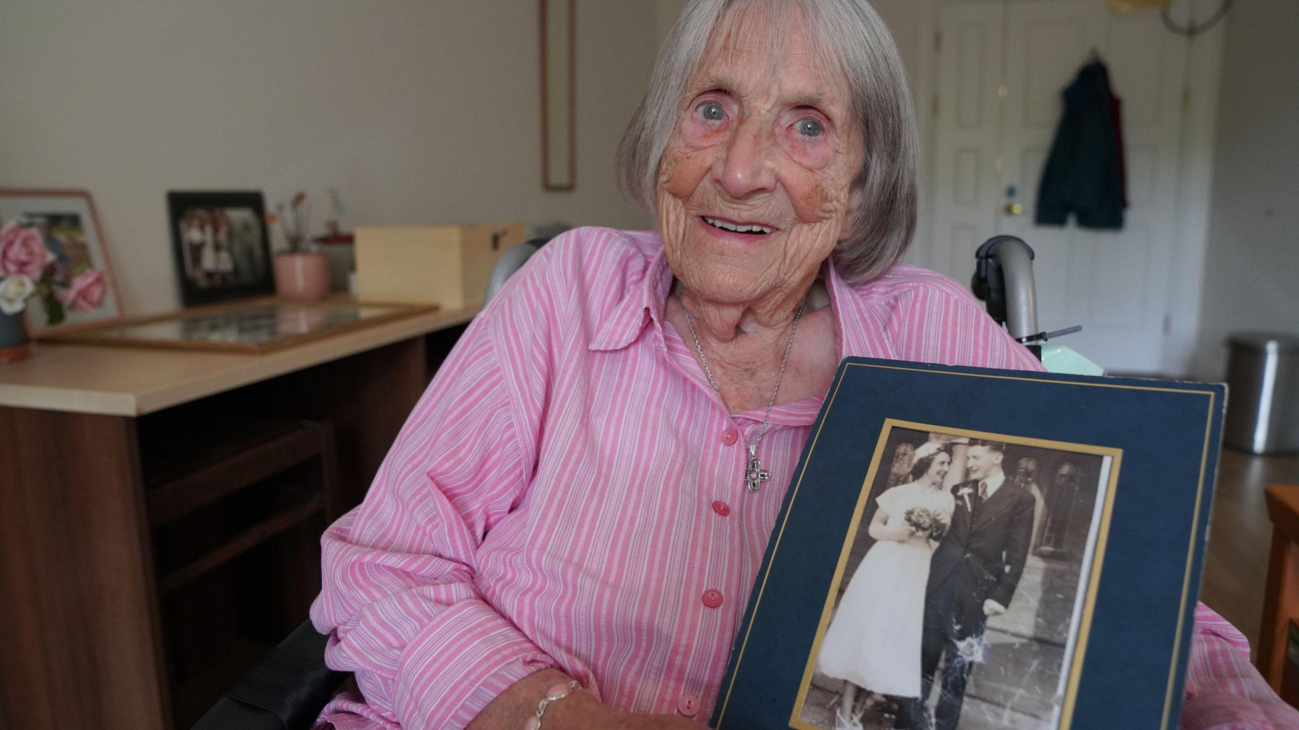an older woman wearing a pink striped shirt sits holding a black and white photograph of her wedding day