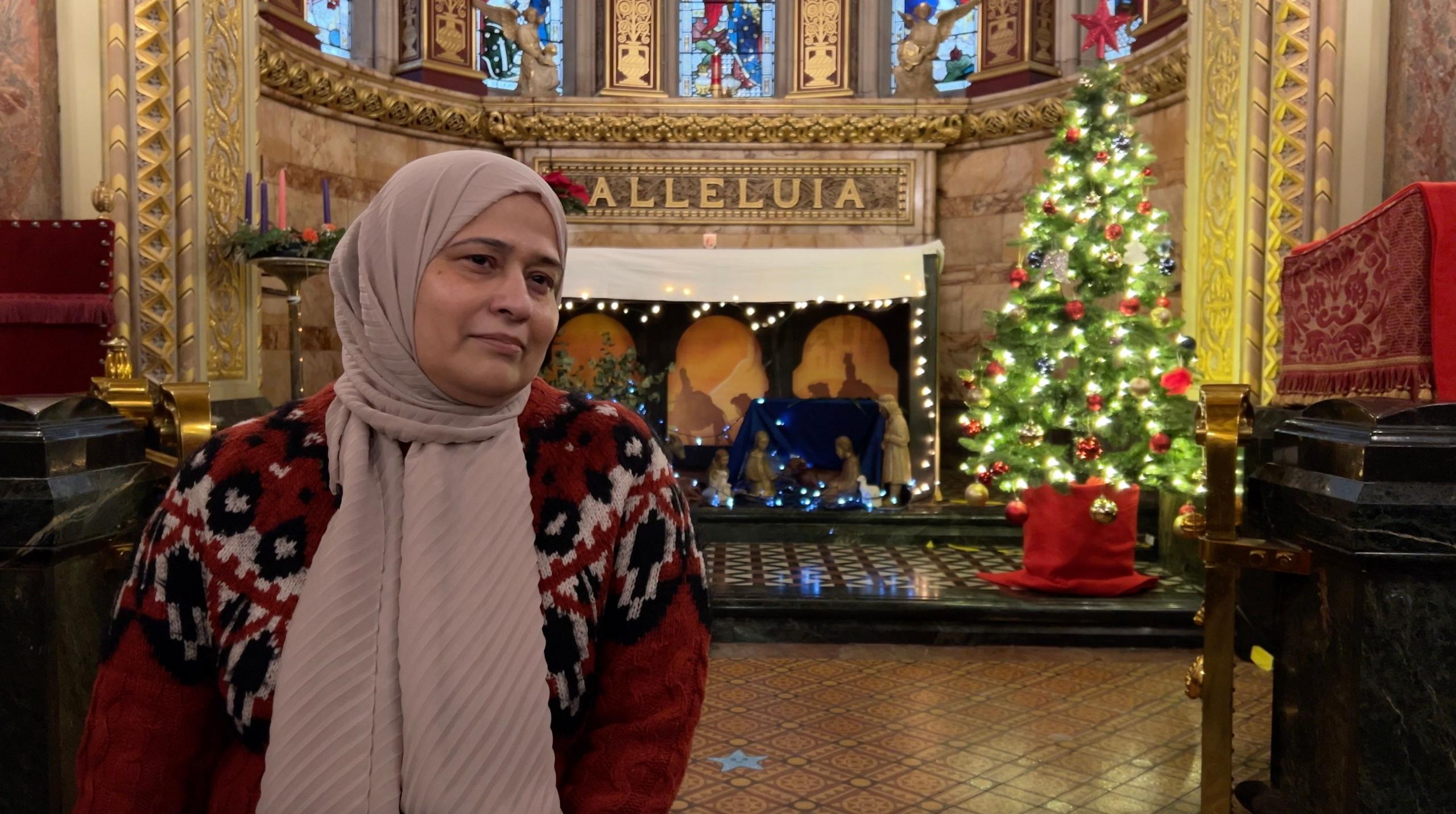 Romana Kazmi, wearing a headscarf and festive red cardigan, in the chapel at Great Ormond Street Hospital, with a lit Christmas tree and nativity scene in the background under the word 'Alleluia'.