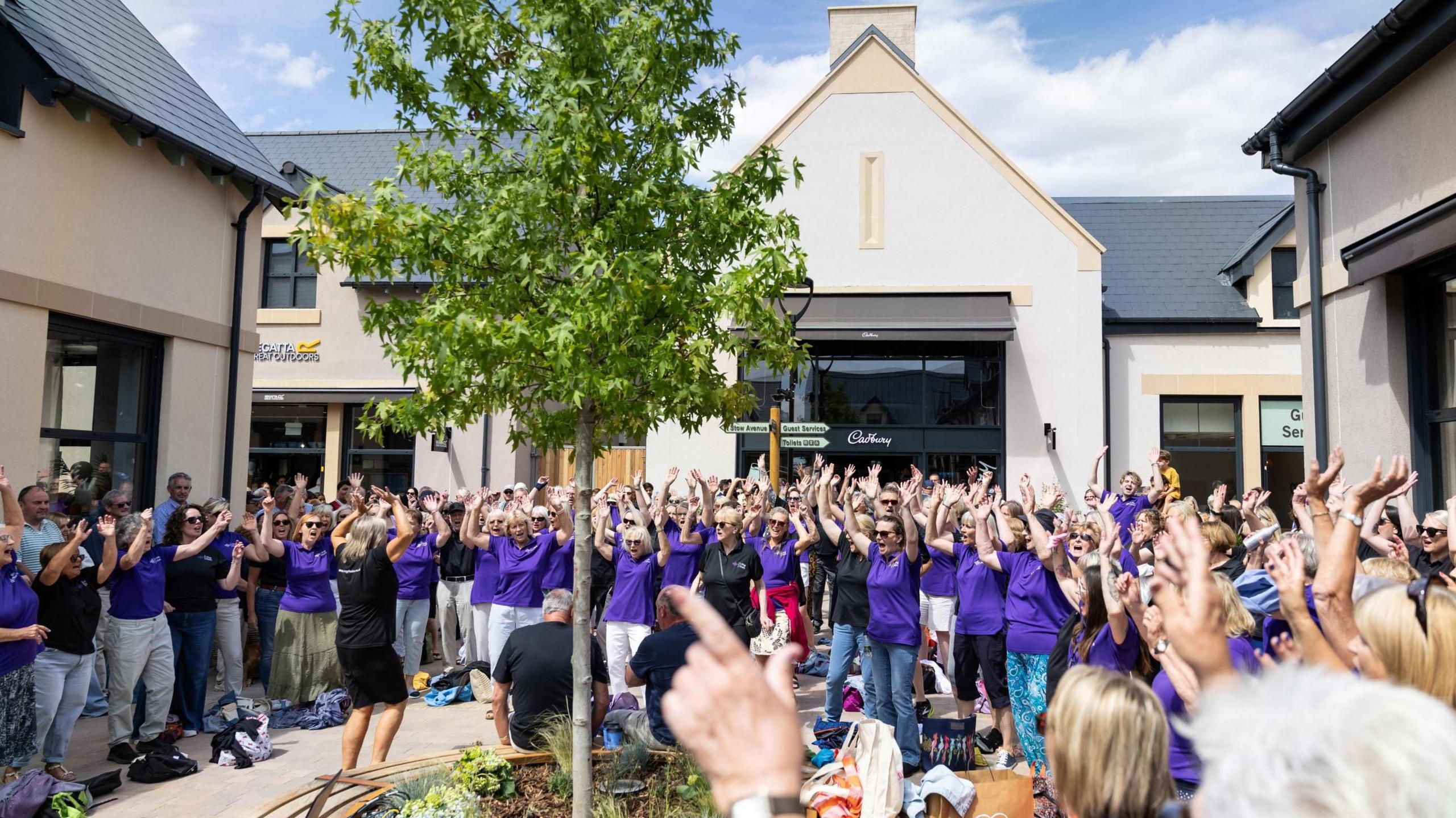 A large group of people wearing purple uniforms at Cotswold Design Outlet. They have their arms raised and appear to be celebrating. 