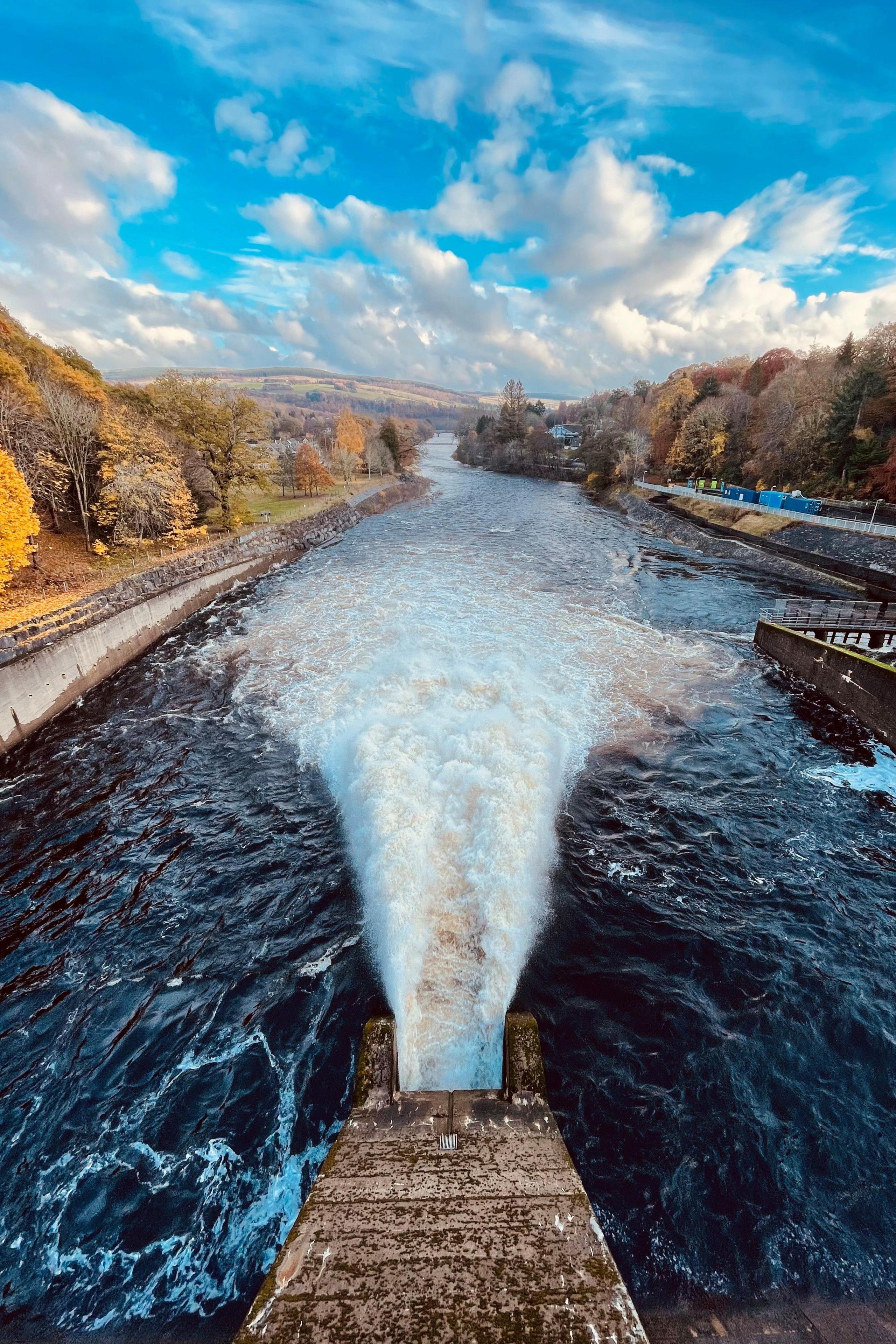 A shot from above of water at the Pitlochry dam, with woodland in the distance. The sky is blue with white clouds.