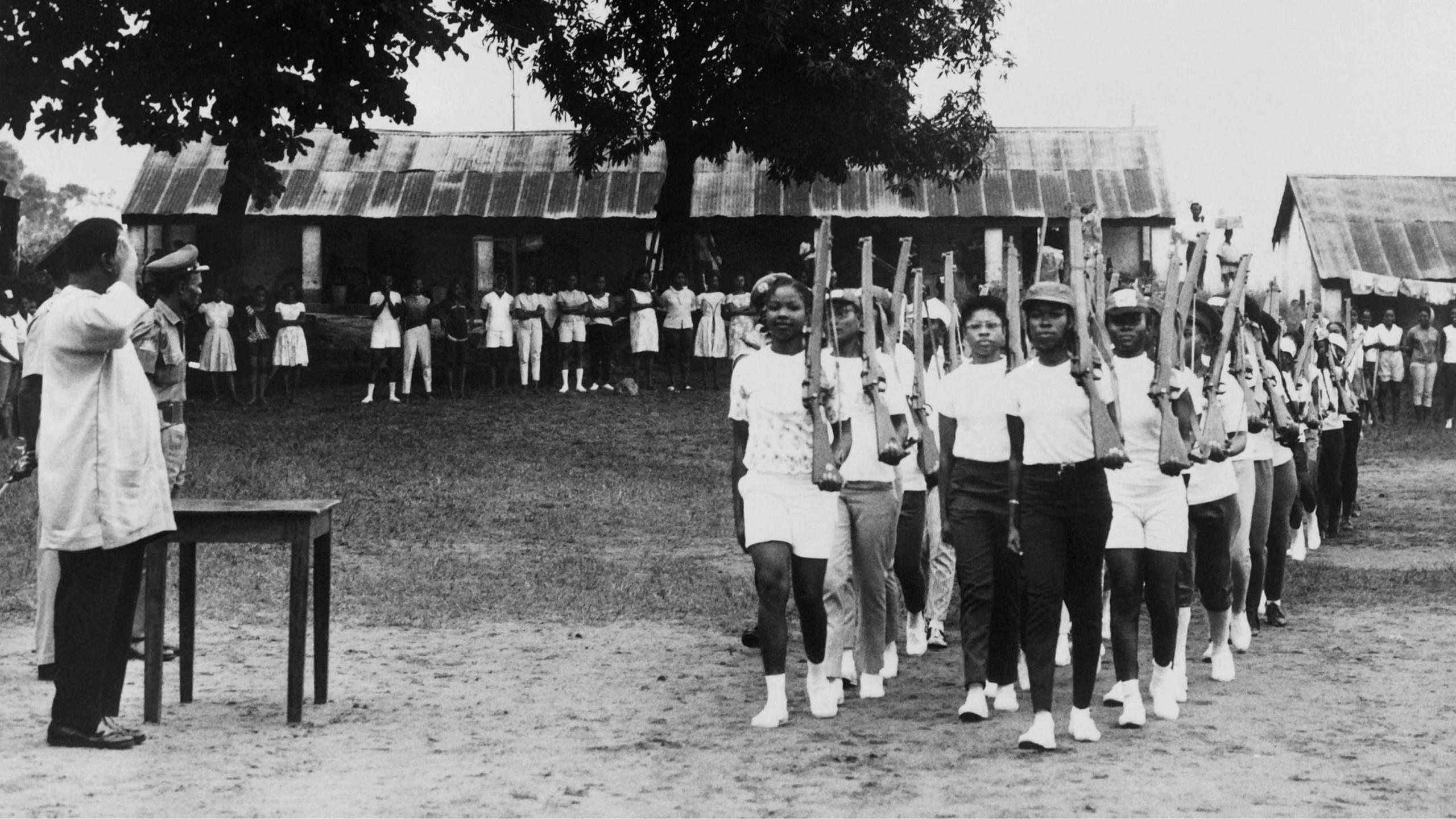 Young female militia of the civil defence - most dressed in white and with white socks and trainers - hold what look like rifles against their shoulders as they parade during military training in Enugu in August 1967. To the side a man in a white coat and dark trousers salutes as two other soldiers look on. It is a black and white archive photo.