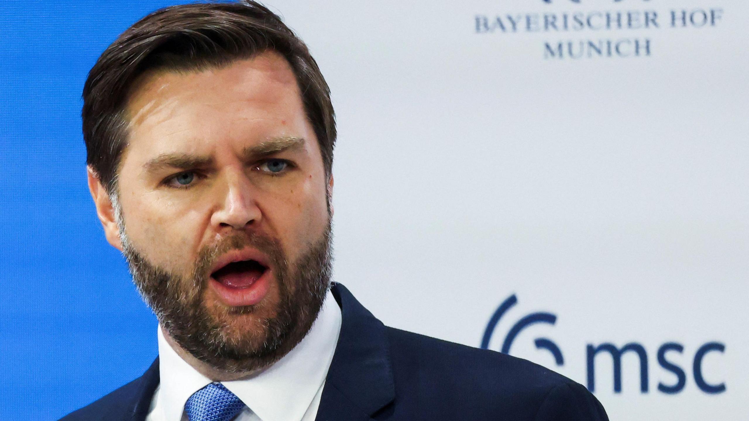 JD Vance - a man with dark hair and a beard, in a blue suit with a blue and American flag lapel pin, giving a speech. 