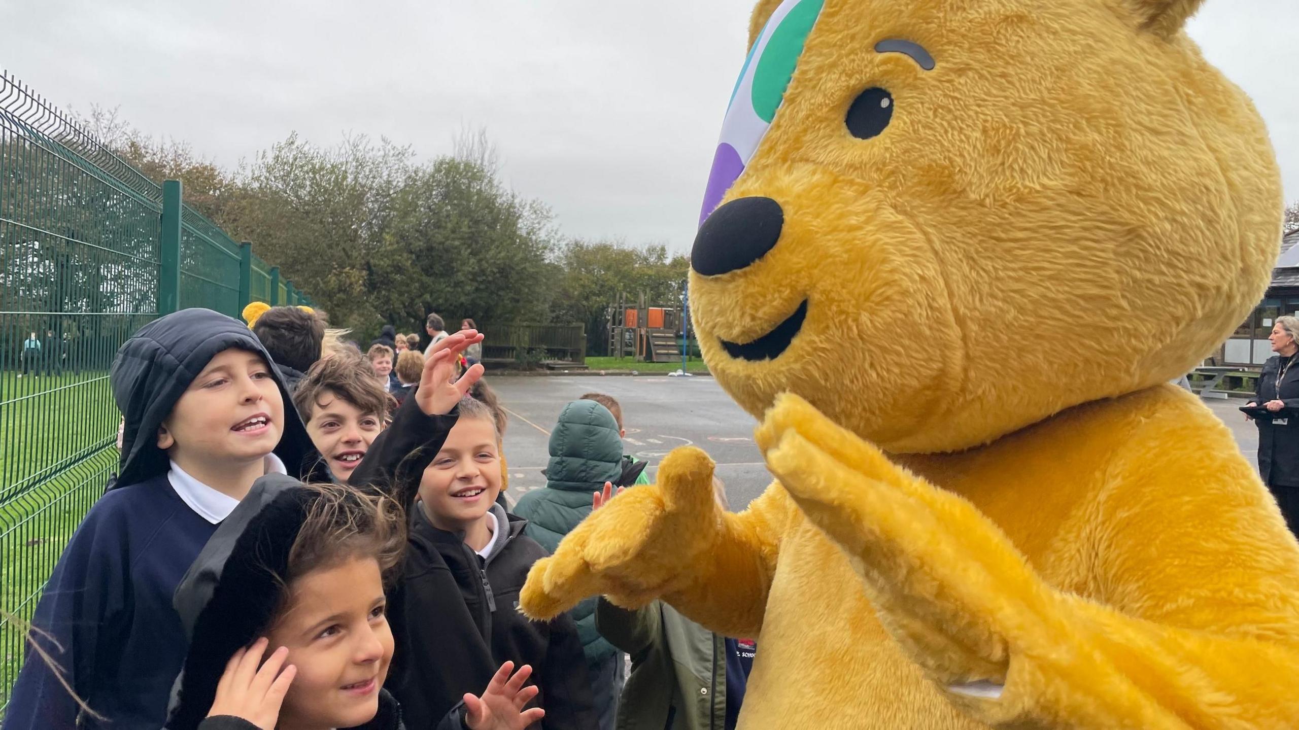 A picture of Pudsey the bear greeting pupils. He is wearing yellow costume. The pupils are pictured on the left of the screen all smiling.