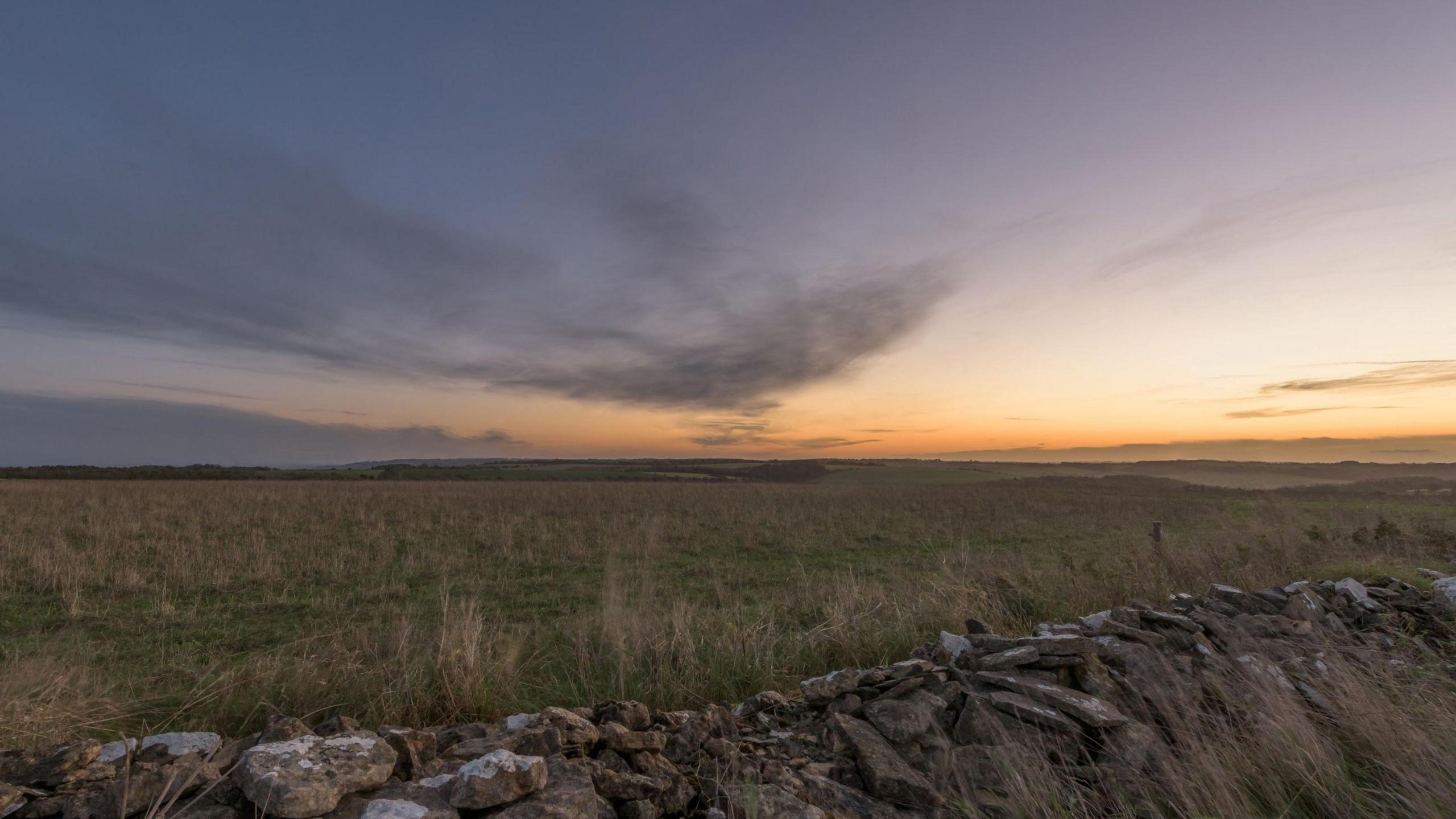 A photo showing a field with a wall at the front and the skies are partly grey with sunshine in the distance