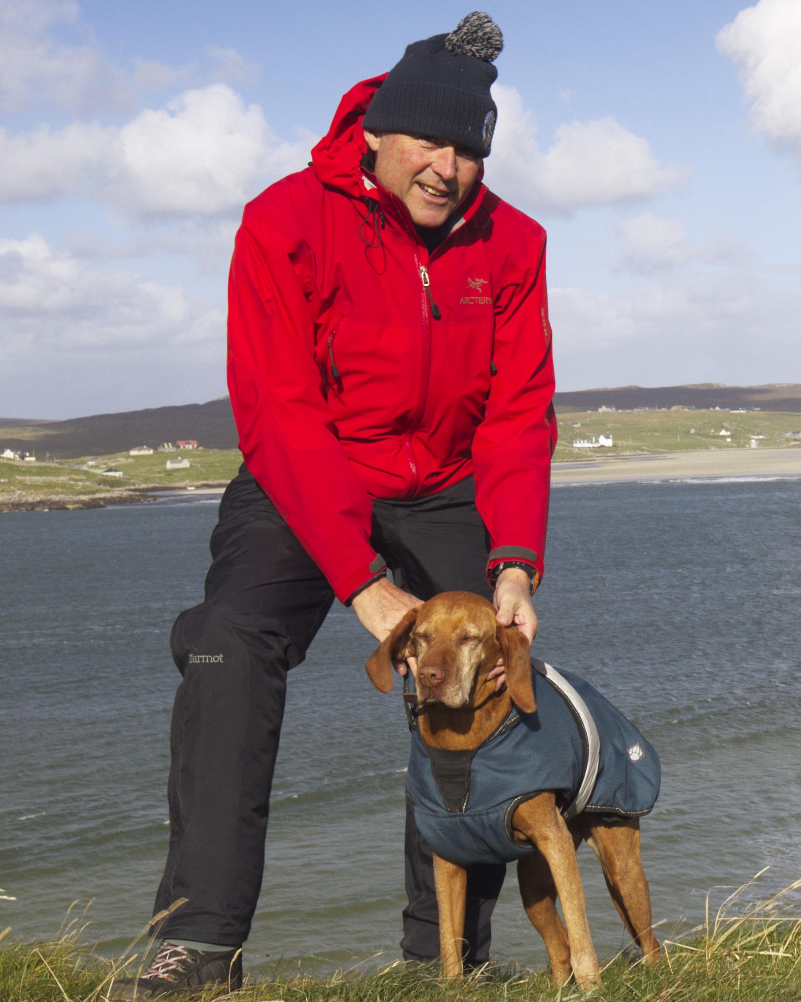 A man in a red waterproof coat, wooly hat and waterproof trousers stands in from of a body of water, with a brown dog beside him