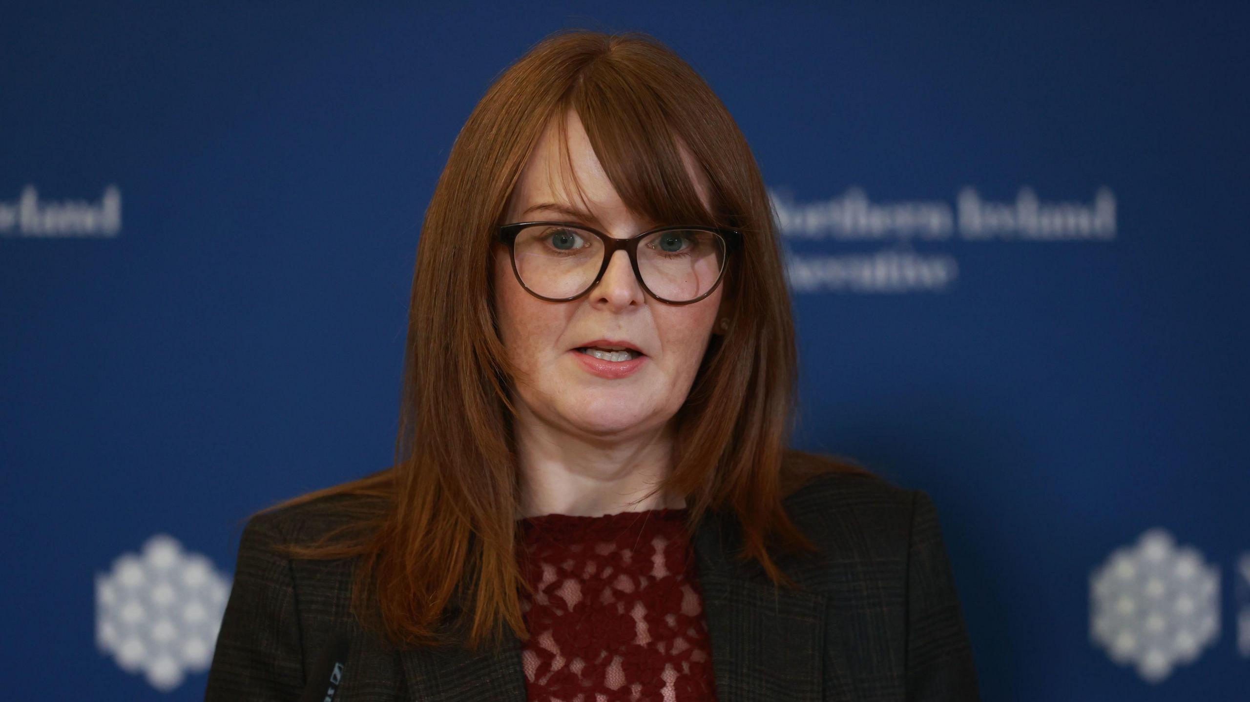 Caoimhe Archibald is standing in front of a background with write writing. She is wearing glasses, a black blazer and burgundy top. 