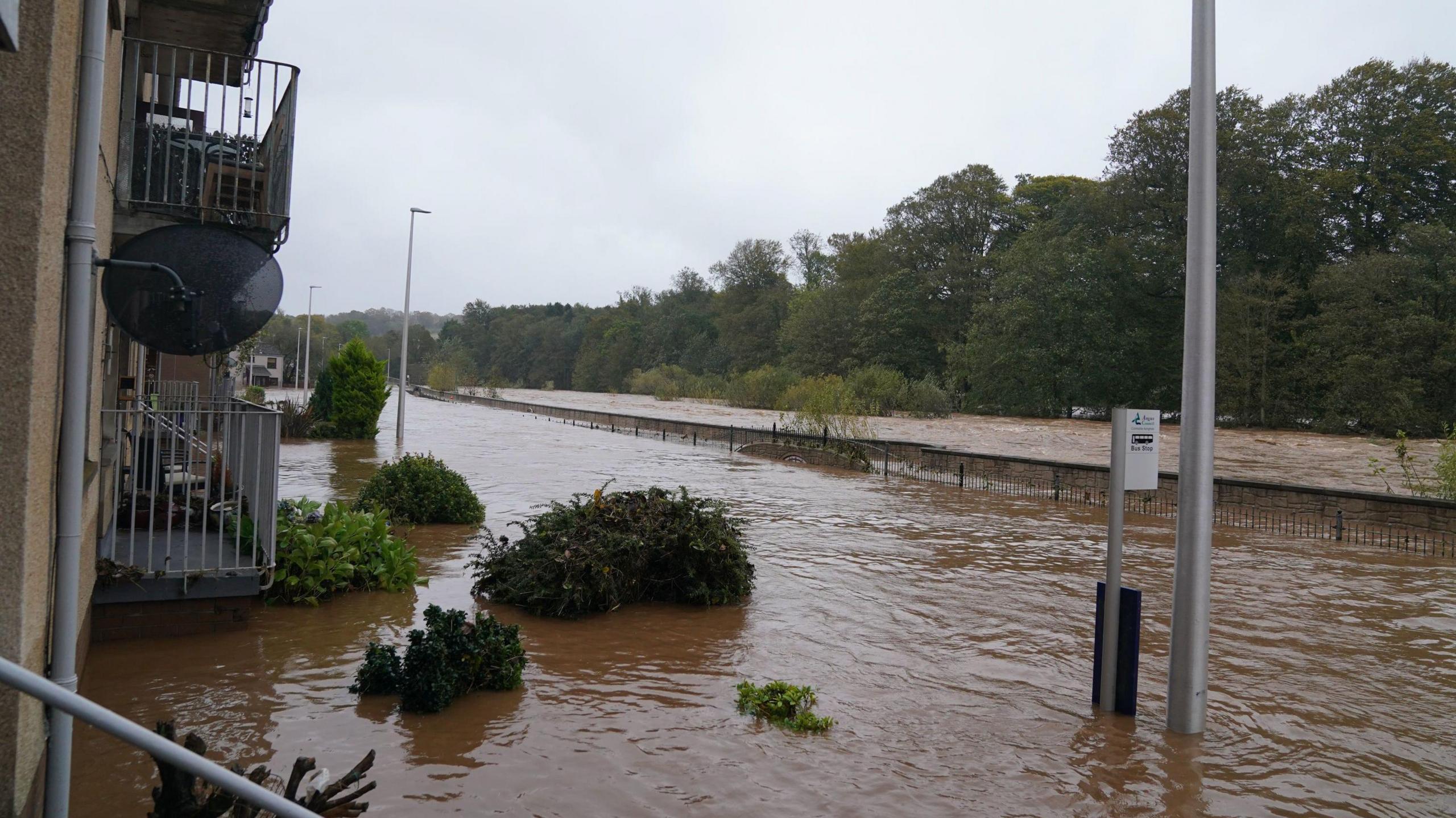 Muddy water reaches what looks like over a metre on a street. Bus stop signs can be seen and the water nearly reaches the heigh of balconies