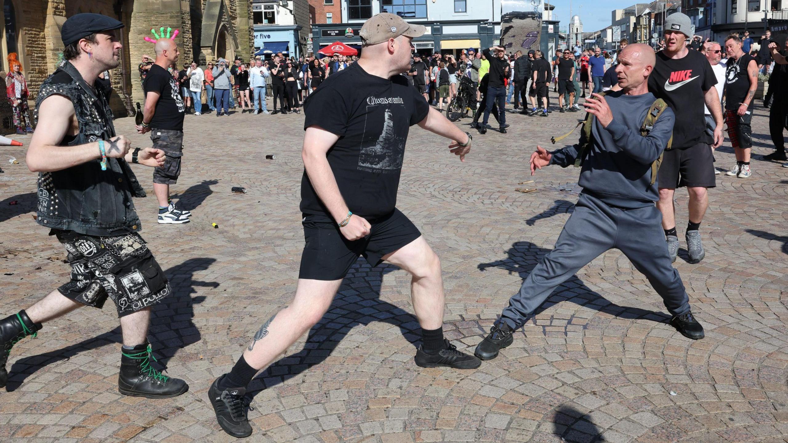 Two men, one in a black T-shirt and shorts and the other in a grey tracksuit, fight outside a church in Blackpool as groups of other protesters stand and watch