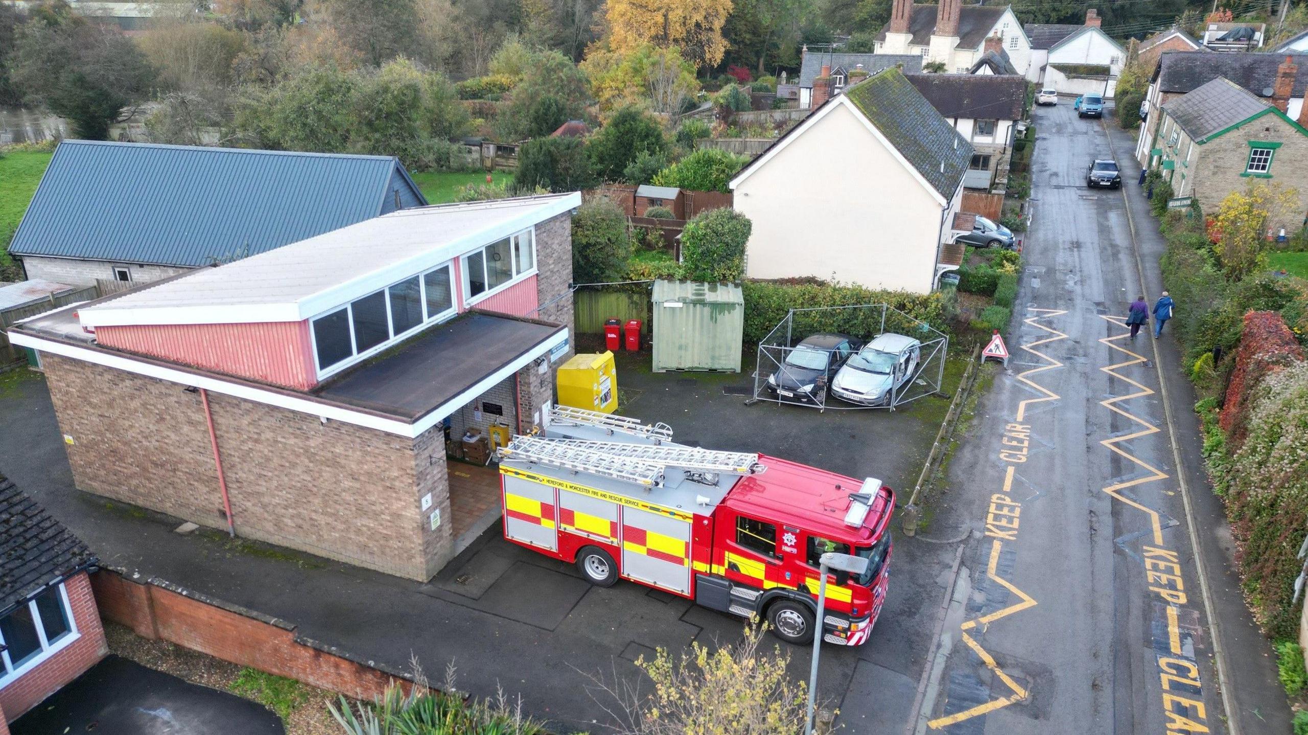 An aerial view of Leintwardine fire station. It is a small one-storey building, with a large open garage and a red fire engine parked in front. There is a road running parallel to the station.