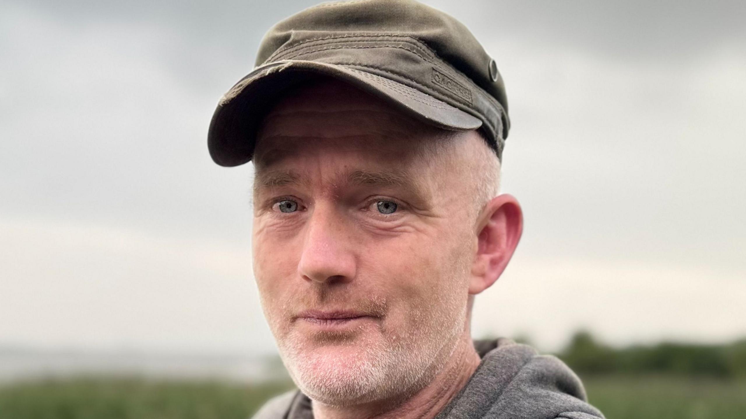 A middle aged white man is wearing a grey hoodie and a brown peaked cap. He has some white stubble on his face. 