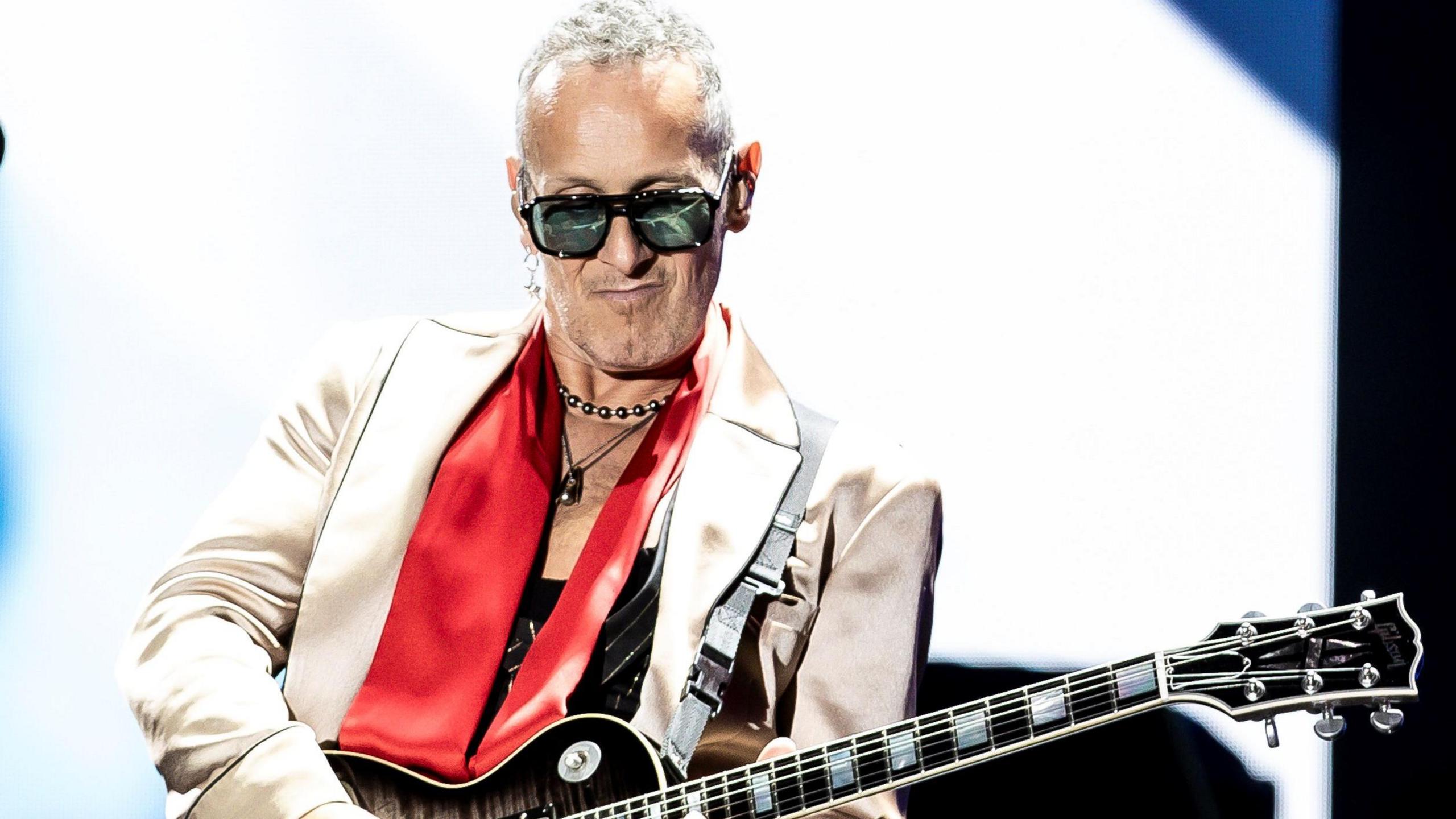 Vivian Campbell is on stage performing. He's playing a guitar. He's wearing sunglasses and a silver jacket with a red scarf and necklaces. White and blue is shining behind him from a large screen.
