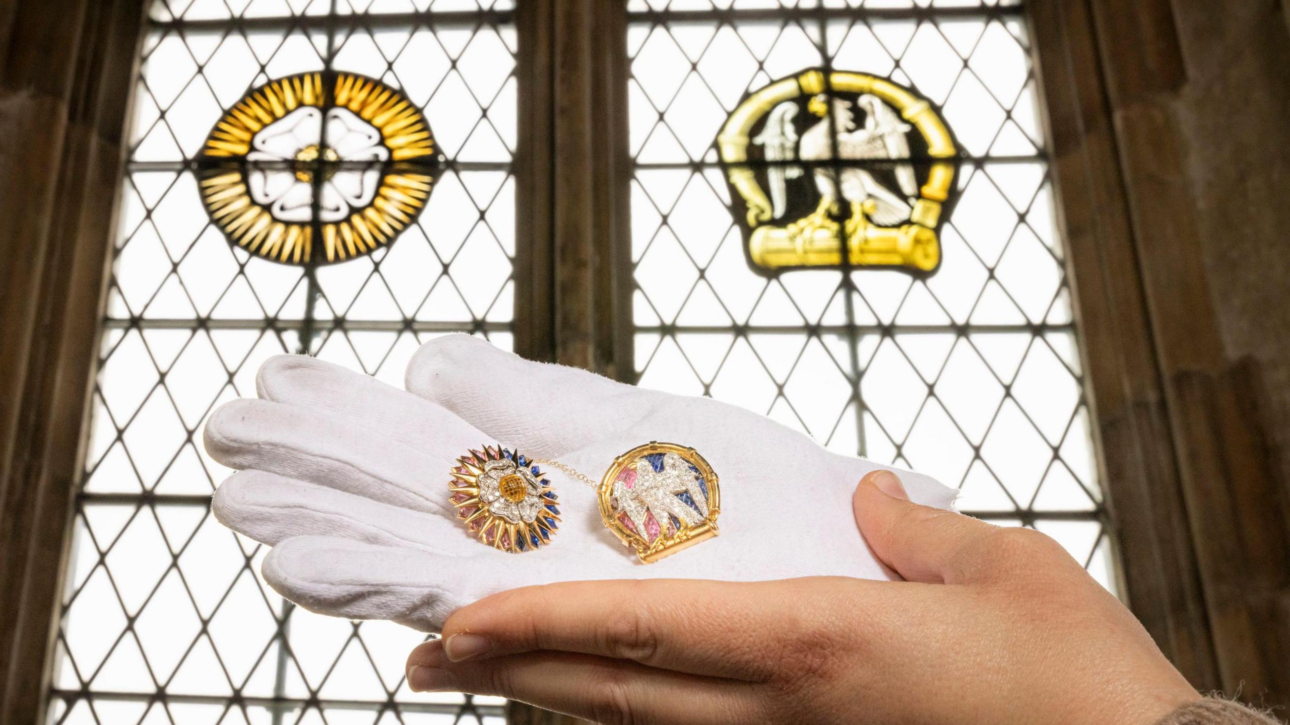 A white gloved hand holds the two brooches up next to the stained glass windows they were inspired by. The brooch on the left features the white Tudor rose and the one on the right is the white falcon.