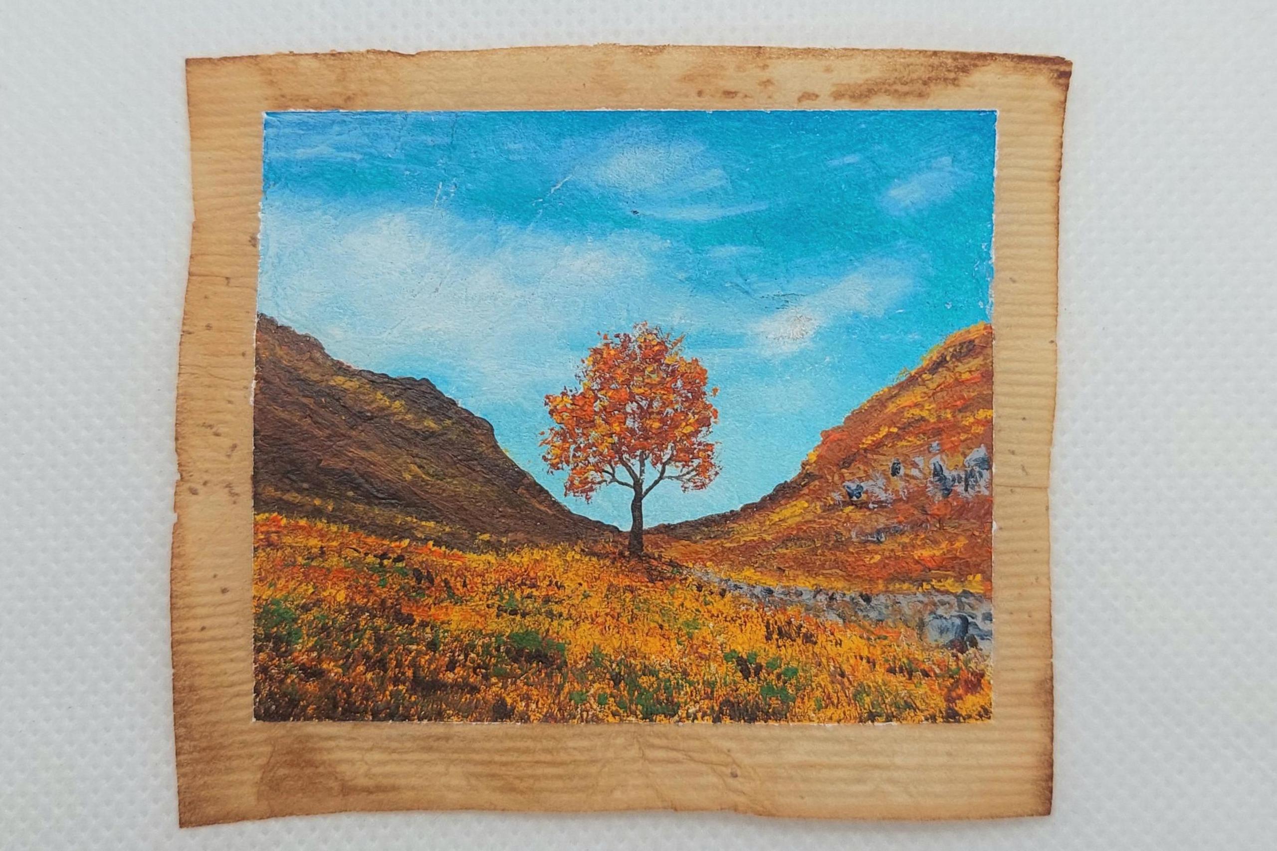 The Sycamore Gap tree on a used teabag painting in an Autumn setting. It has orange leaves and the field around it is in Autumnal colours.