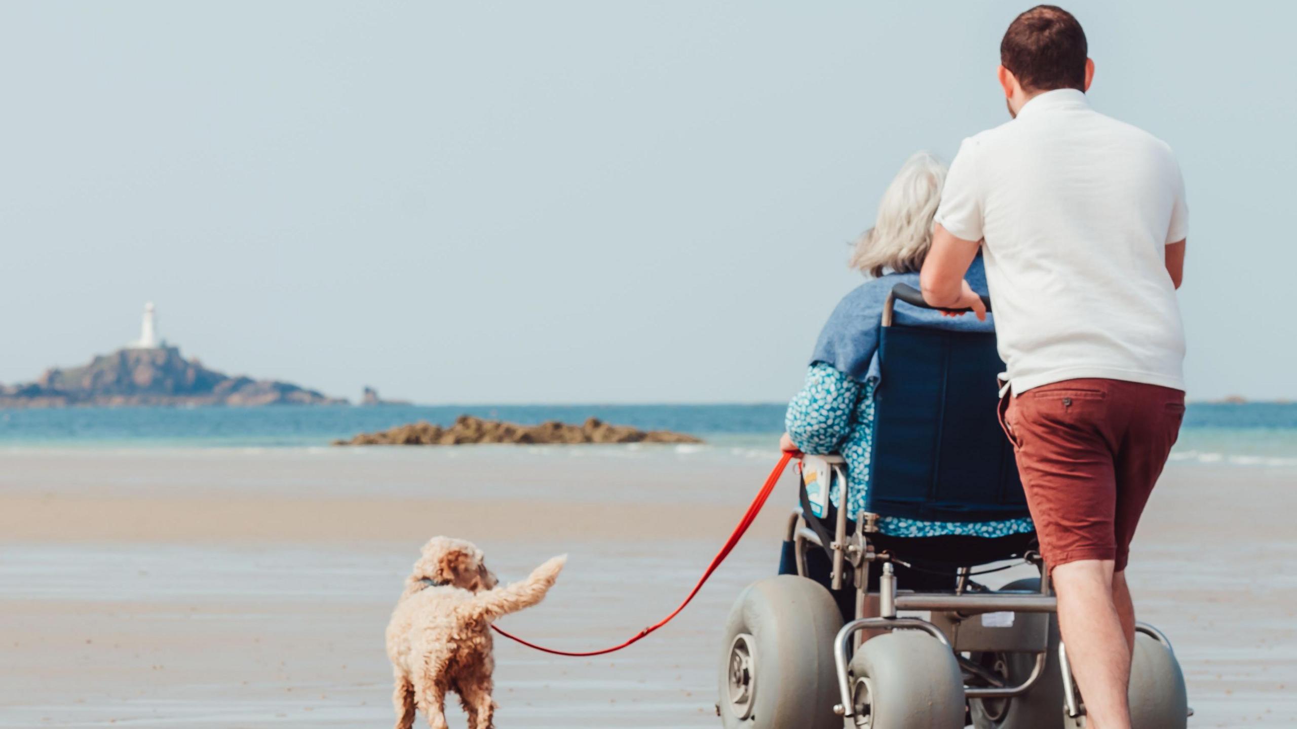 A man his pictured pushing a lady in one of the beach wheelchairs in jersey on a beach. They are also with a golden colour dog.