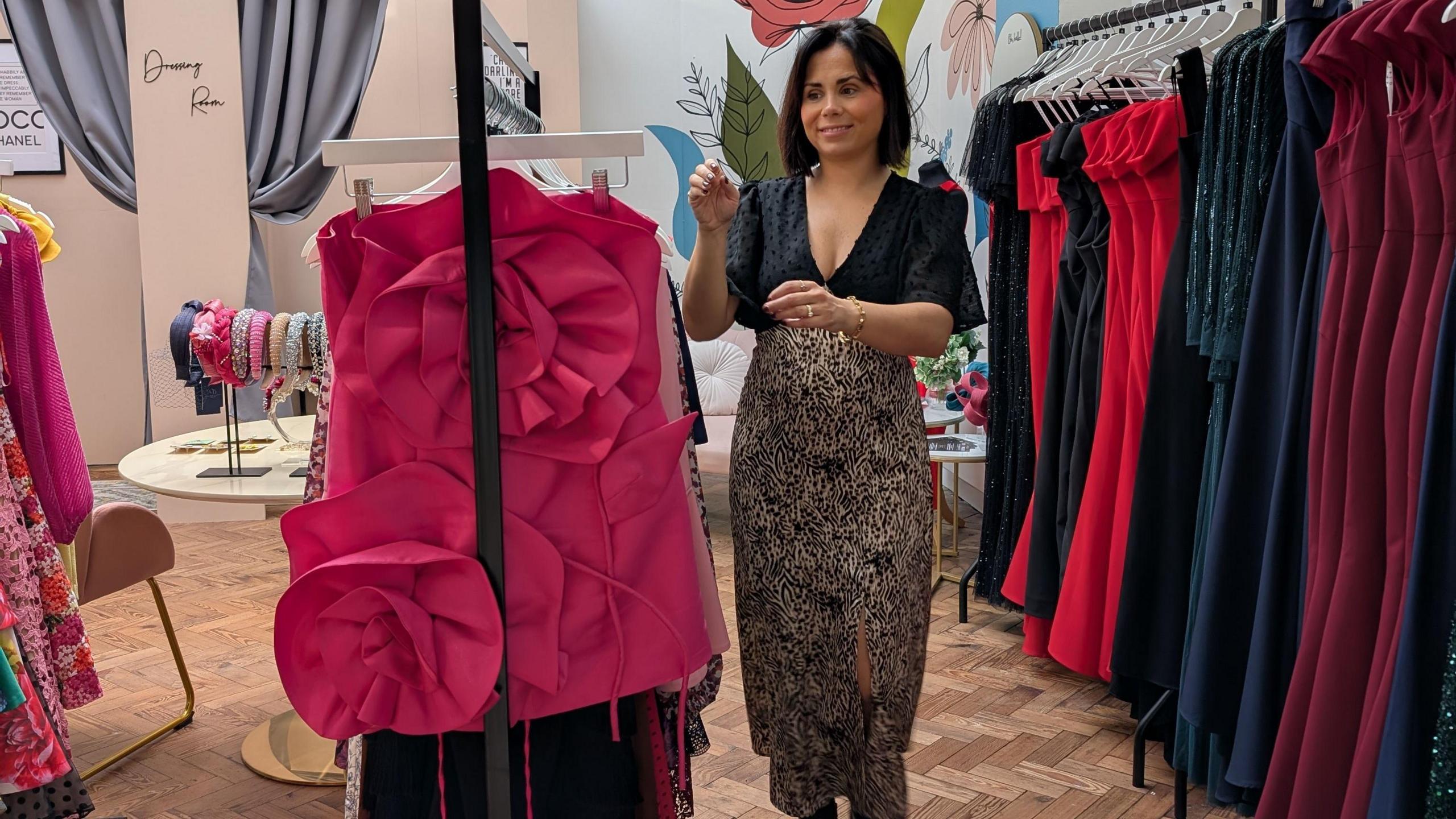 A woman wearing a black top and skirt stands in a shop. Rows of dresses and partywear surround her.