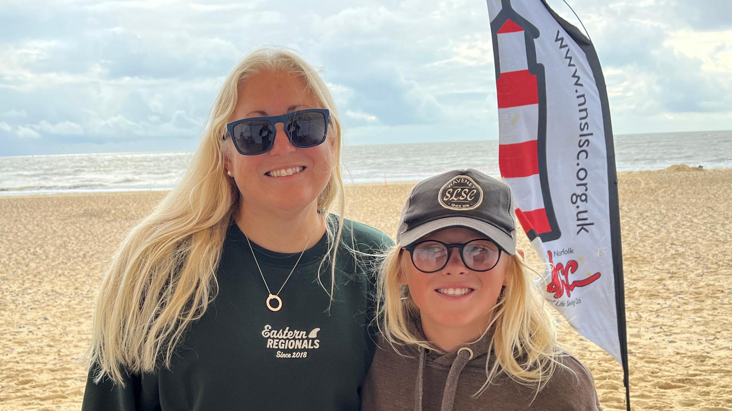 Caroline Harker smiles at the camera on the beach and stands alongside on of her sons. They both have long blonde hair. Caroline wears sunglasses and a dark green jumper. Her son wears a grey cap and dark grey hoodie.