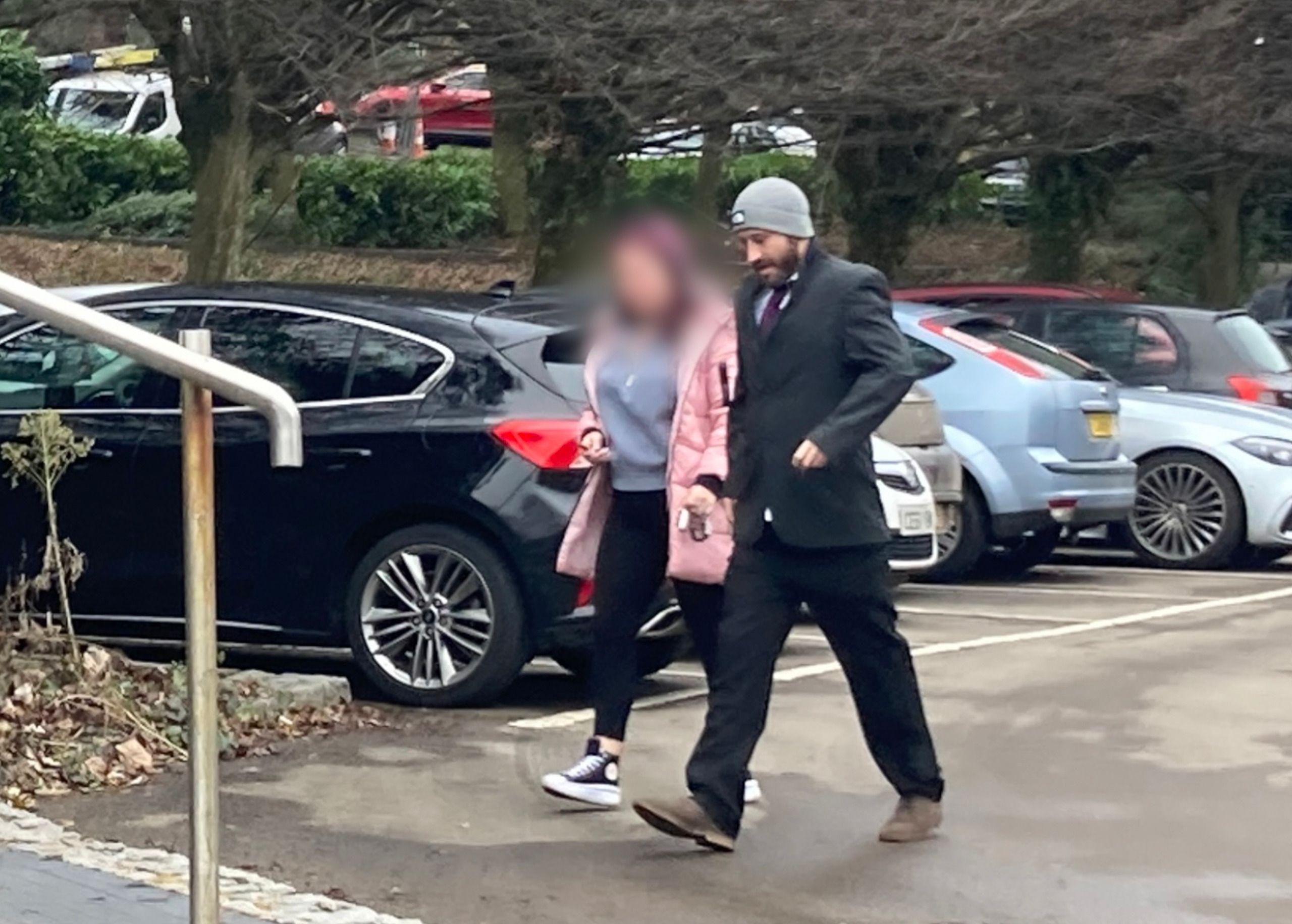 Justin Allison dressed in a black suit and grey hat walking through a car park with a woman in a pink coat.