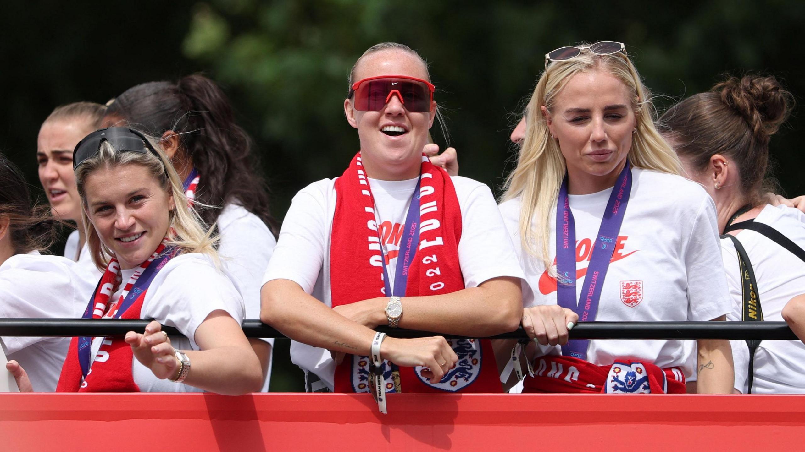 Three england footballers, wearing white tshirts and medals on purple lanyards around their neck, are looking over a black metal railing. 
